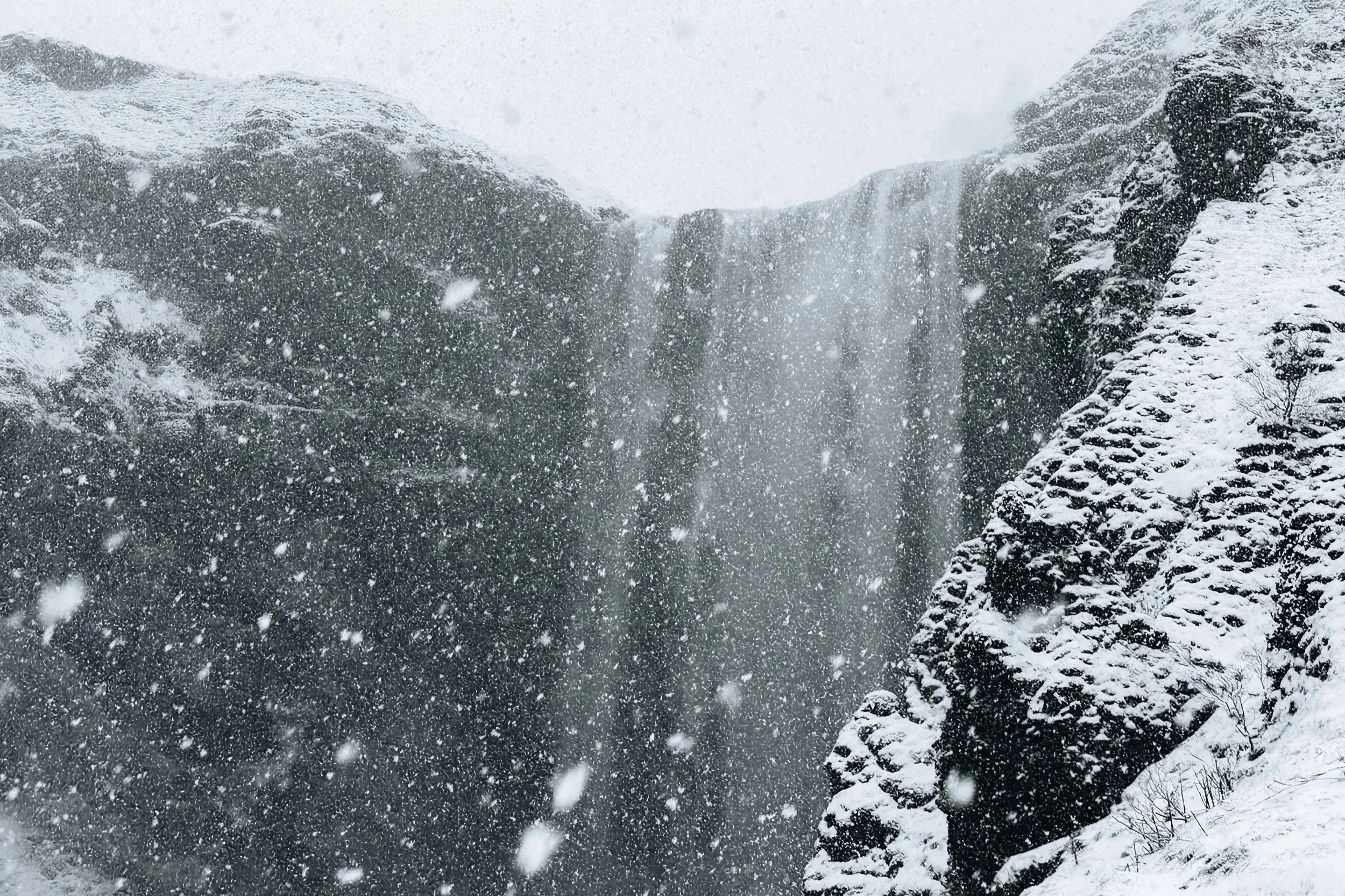 Snow falling at a waterfall surrounded by snow-covered rocks and cliffs.