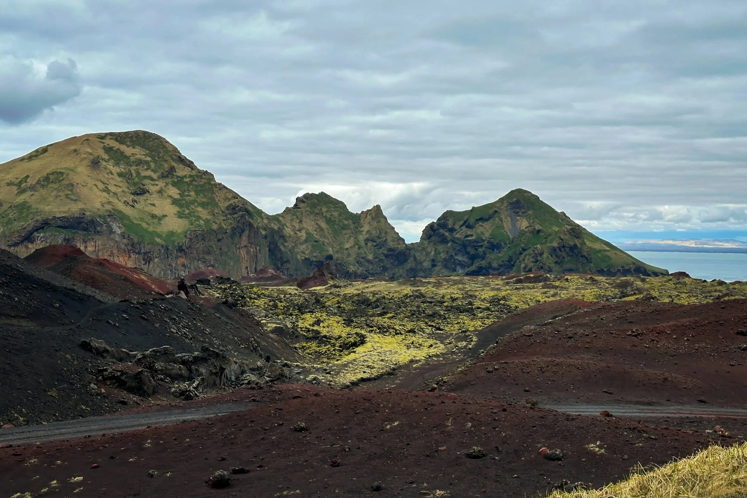 View of volcanic landscape with dark reddish-brown soil and green moss-covered rocky mountains in the background under a cloudy sky, with a distant body of water on the horizon.