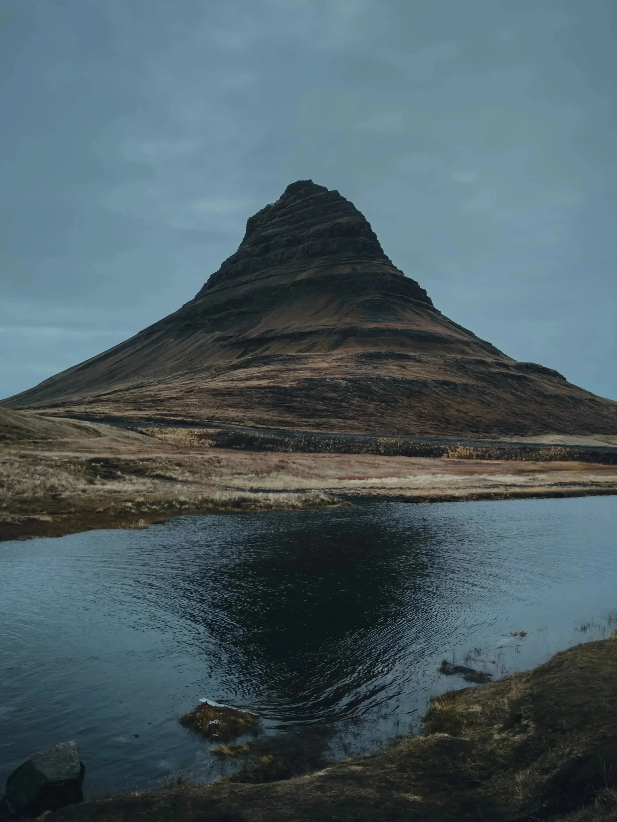 A mountain with a conical shape reflecting in a body of water, surrounded by a grassy landscape under a cloudy sky.
