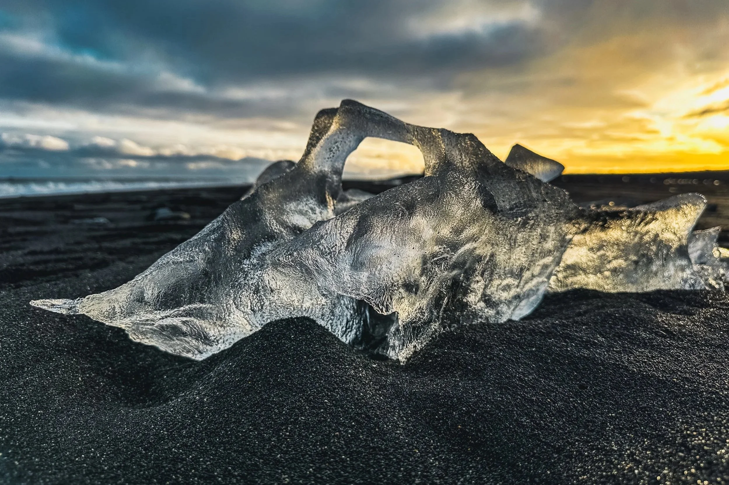 A large, clear ice fragment on black sand beach during sunset with cloudy sky.