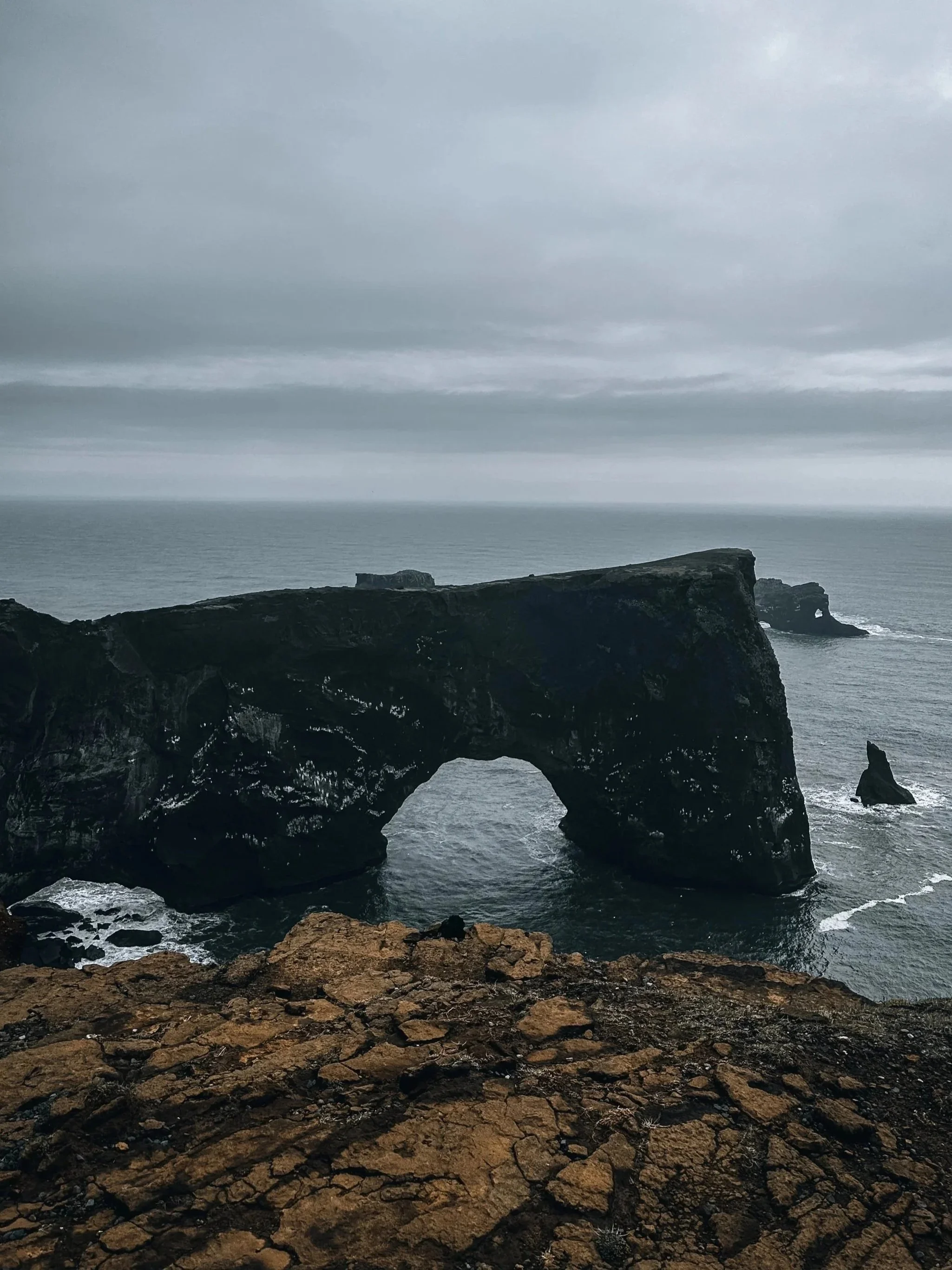 A dark, rocky coastal landscape with a natural rock arch formation over the water under a cloudy sky.