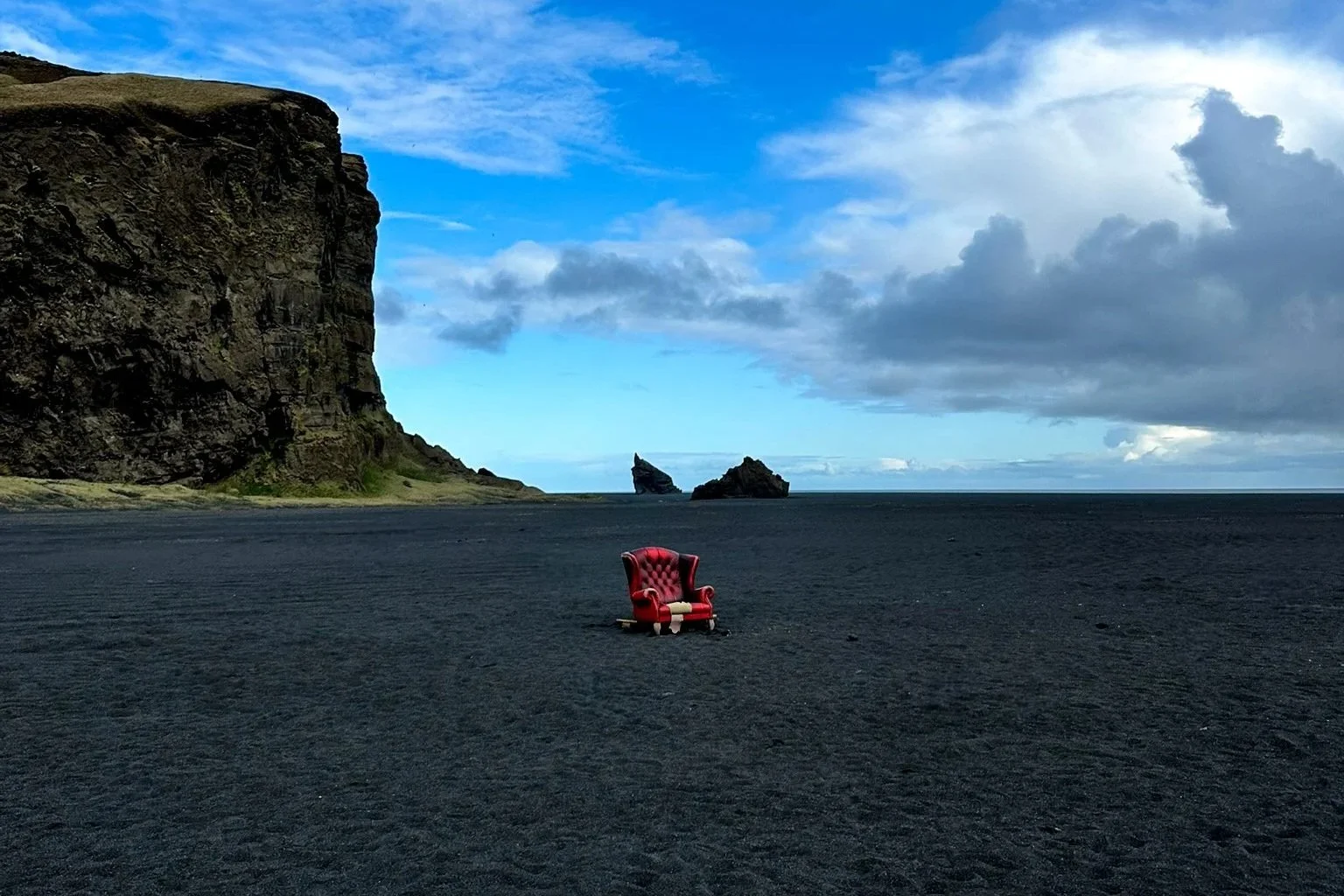 A red velvet armchair on black sand beach with cliffs and ocean in the background, under a partly cloudy sky.