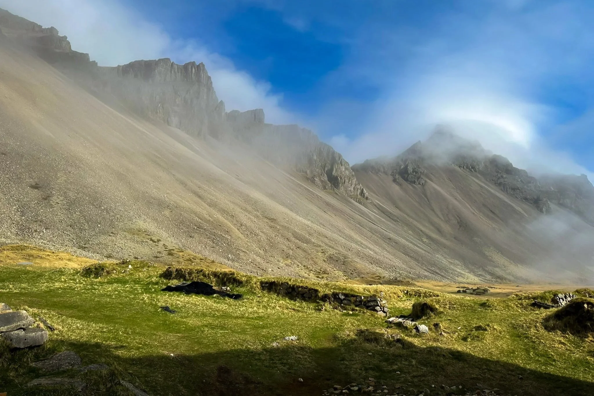 A misty mountain landscape featuring tall rocky mountains with clouds around their peaks, a grassy field with rocks in the foreground, and a blue sky with some clouds.