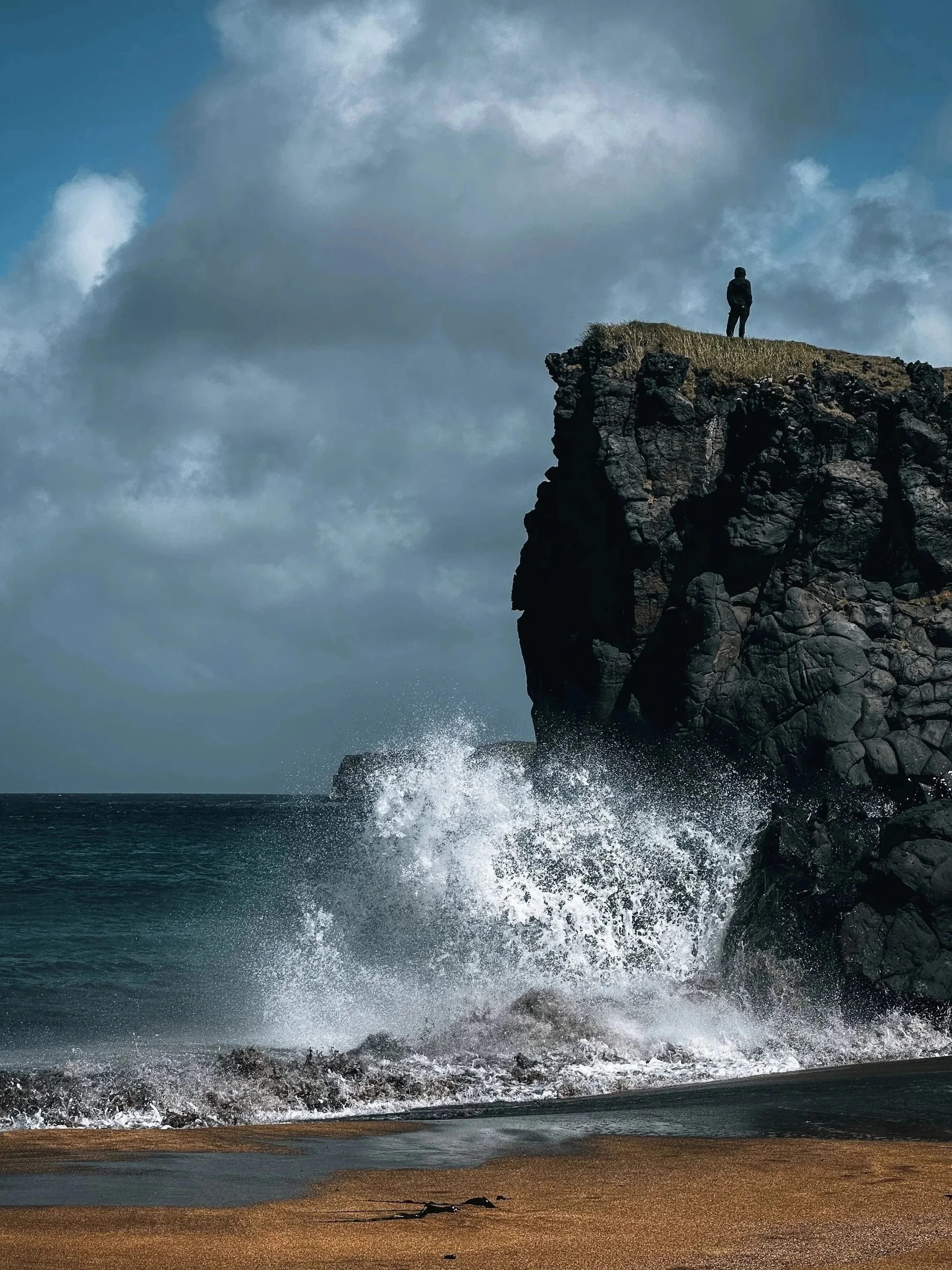 A person standing on a grassy cliff overlooking the ocean as waves crash against the rocks below under a cloudy sky.