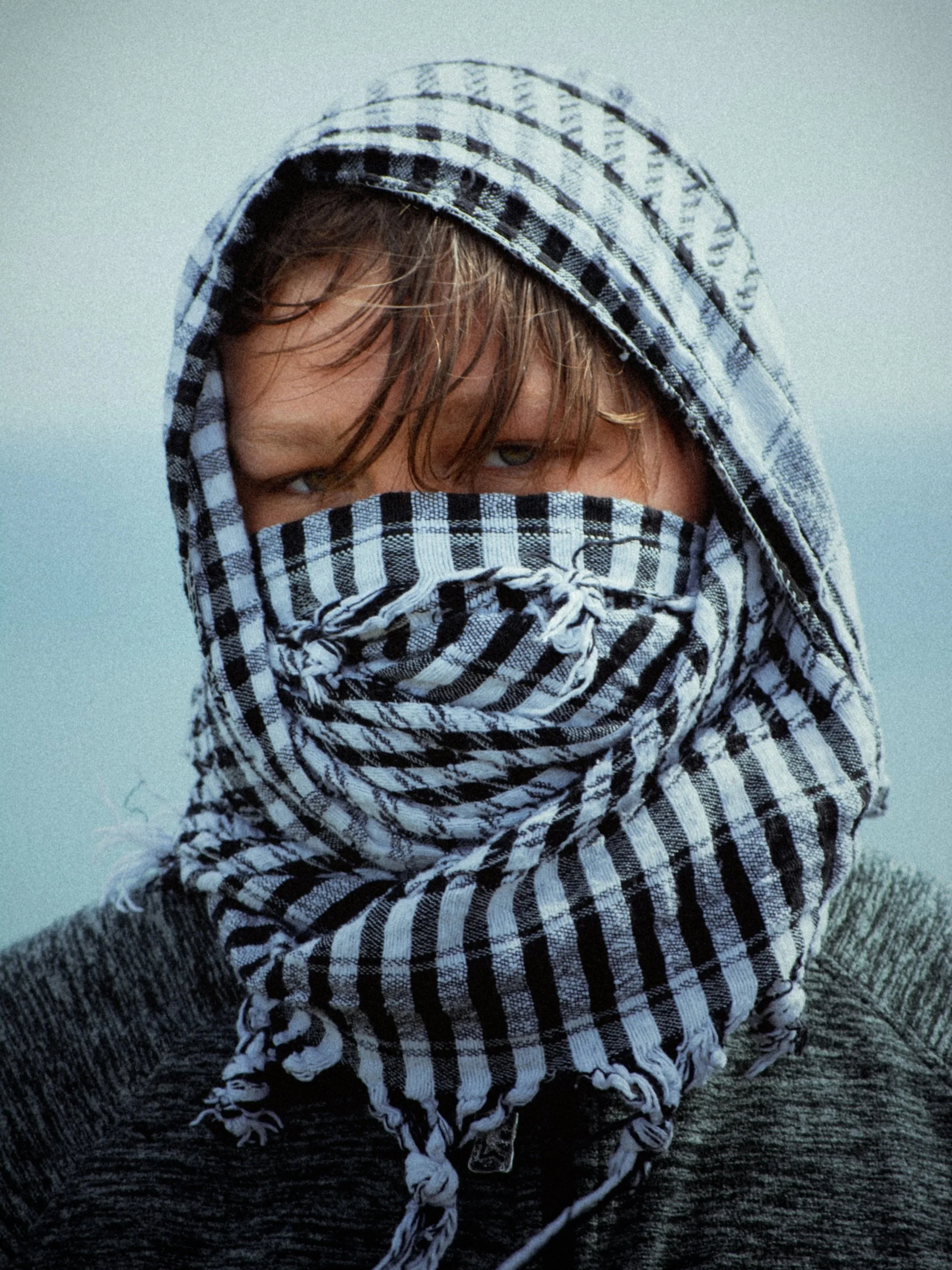 Person with wet hair wearing a black and white checkered scarf over face and head, gray sweater, against a cloudy sky.