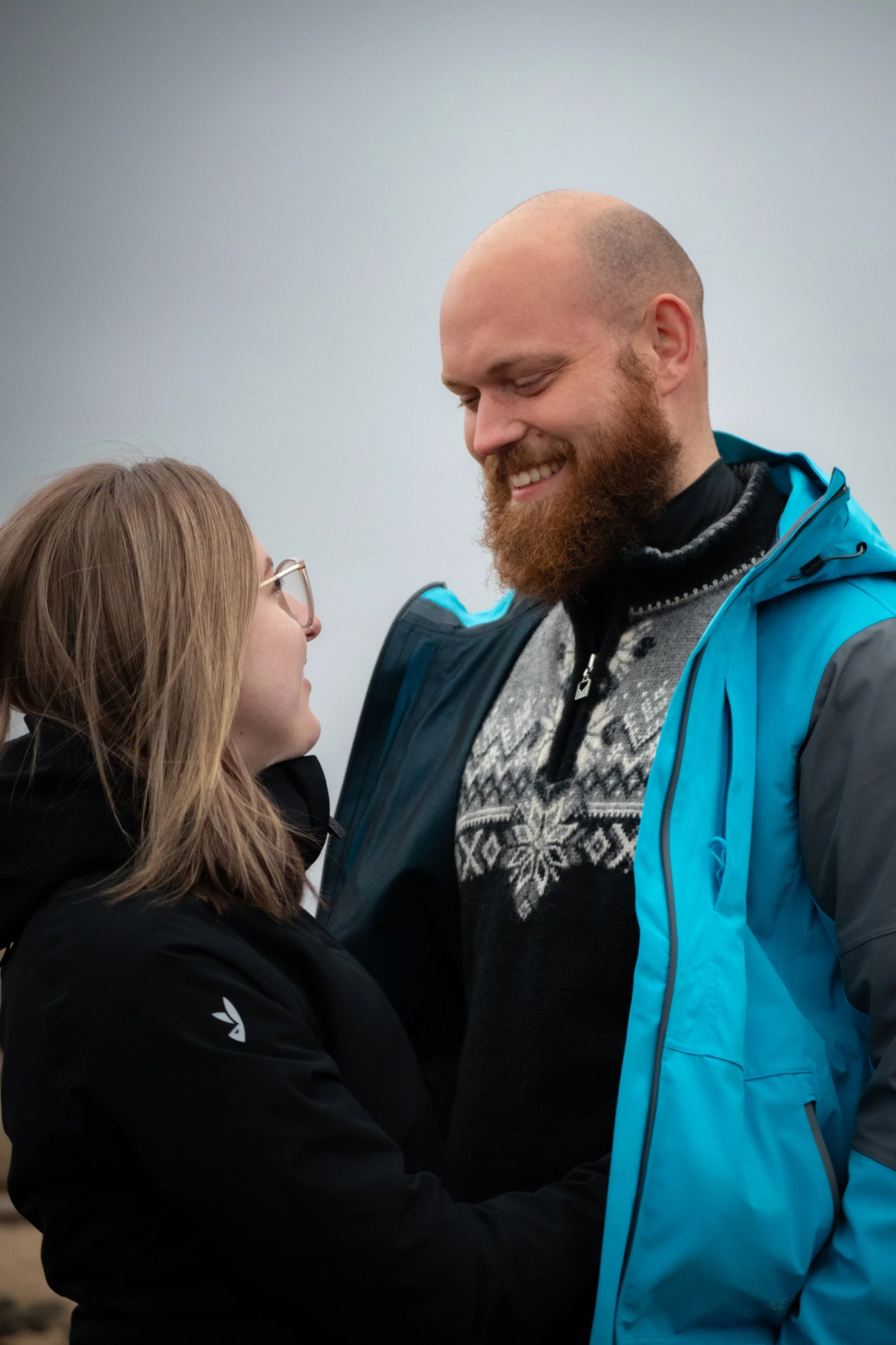 A woman and a man are facing each other, smiling, indoors against a plain background. The woman has long brown hair, glasses, and is wearing a black jacket. The man is bald, has a red beard, and is wearing a blue jacket over a patterned sweater.