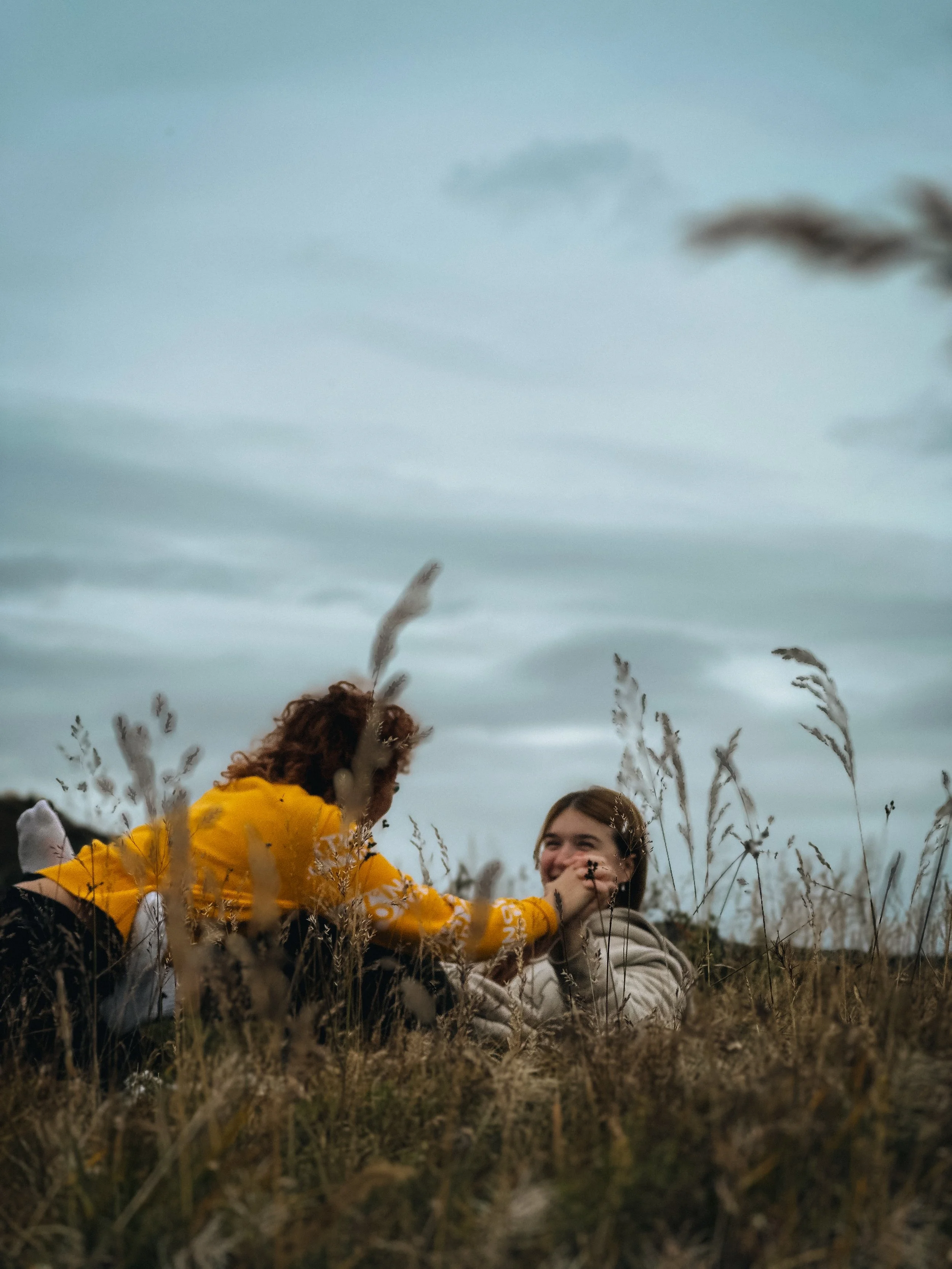 Two people sitting in tall dry grass outdoors, one wearing a yellow hoodie and the other in a beige jacket, smiling and interacting under a cloudy sky.