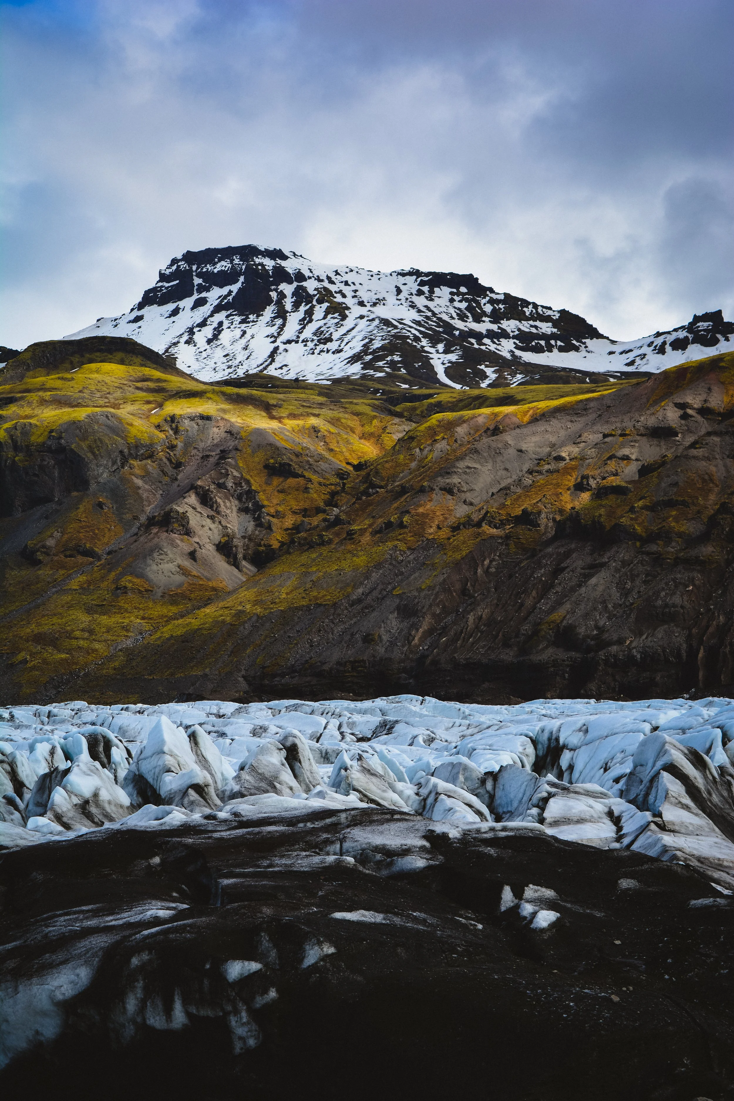 Snow-covered mountain with green and brown rocky slopes, icy glacier in the foreground, and a cloudy sky above.