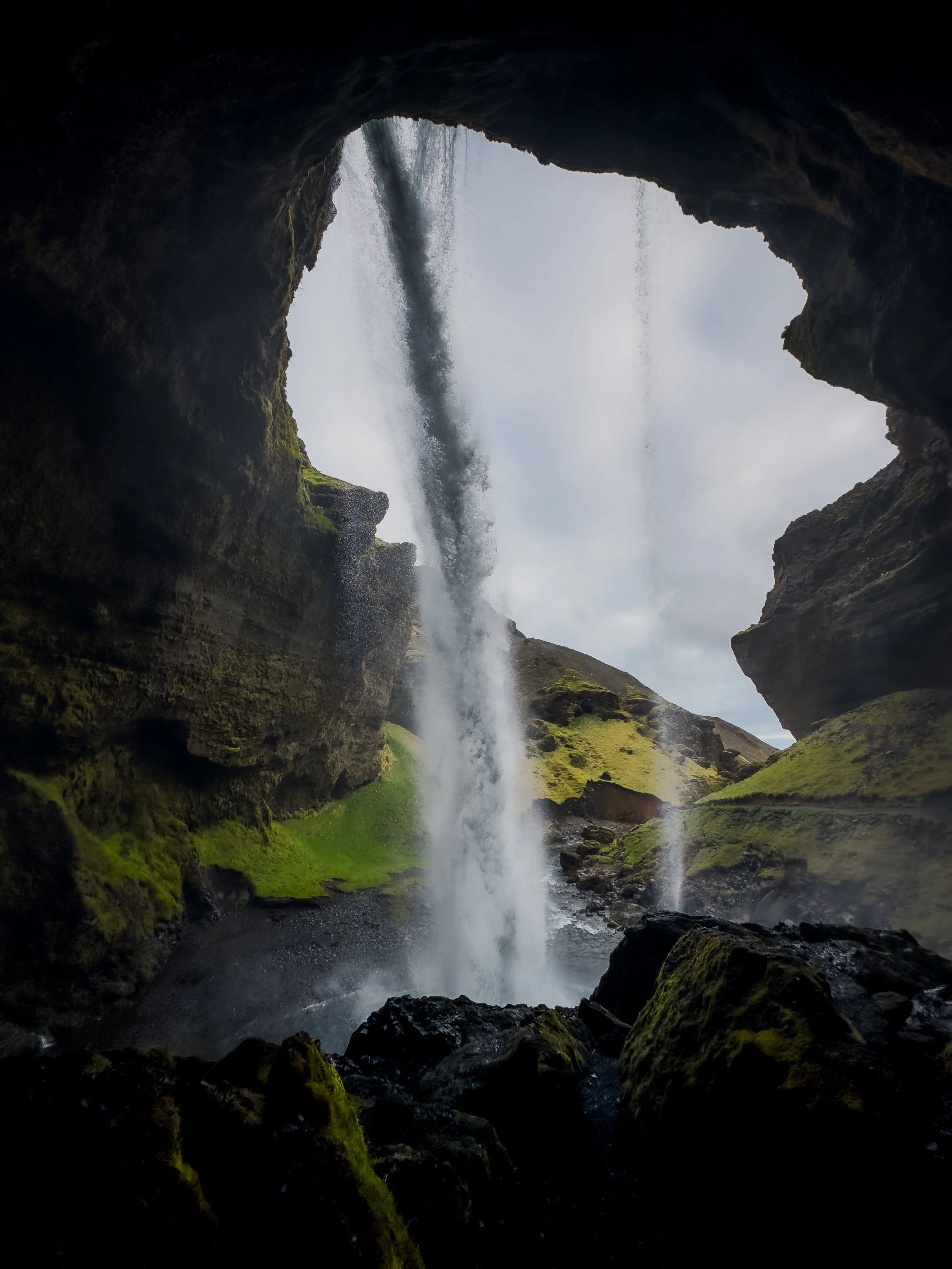 View from inside a cave looking out at a waterfall cascading down a moss-covered cliff into a stream below.
