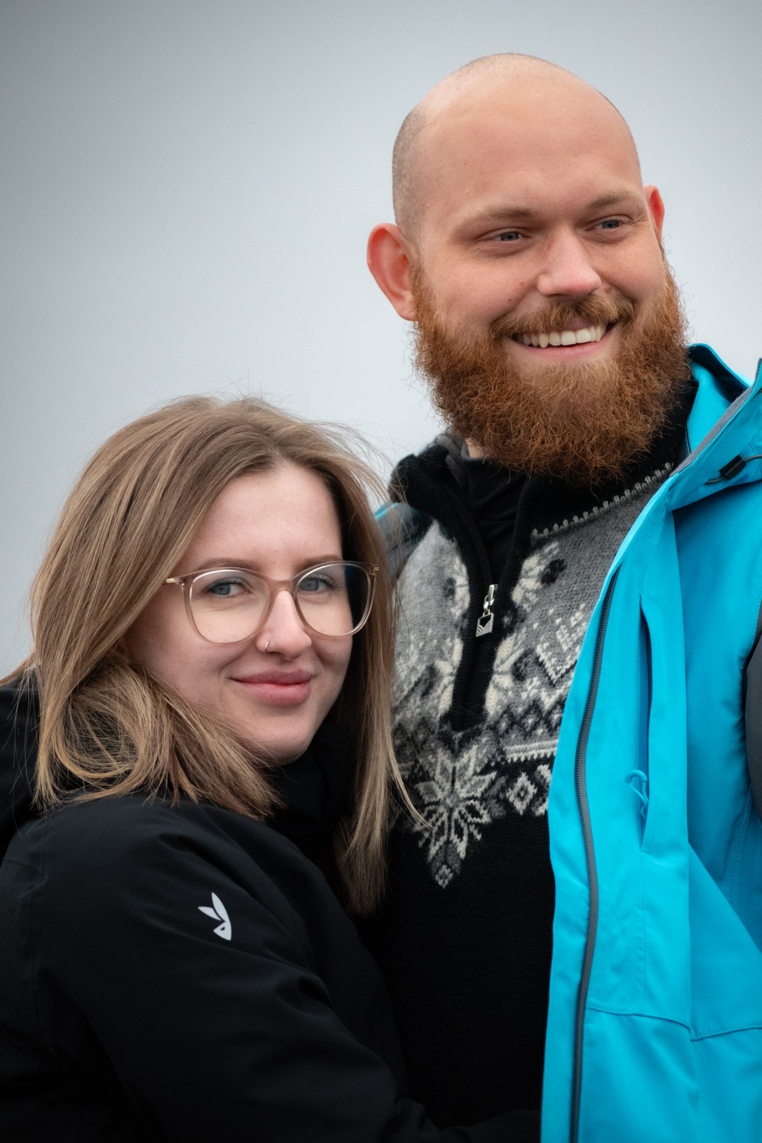 A woman with long, light brown hair, glasses, and a nose piercing smiling, and a man with a beard and a shaved head smiling, standing close together outdoors against a cloudy sky.