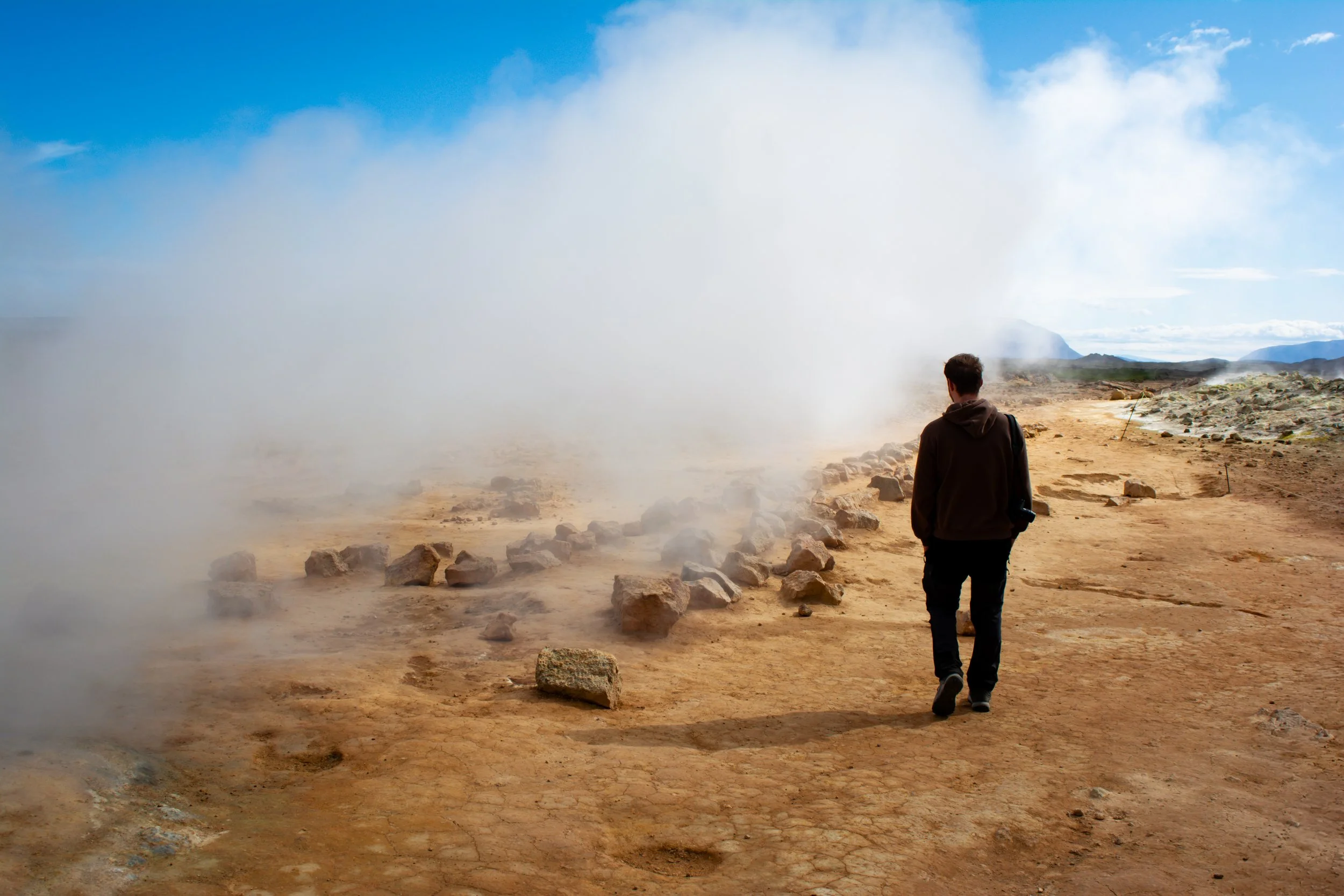 A person walking on a rugged dirt trail through a geothermal area with steam rising from the ground and a mountainous landscape in the distance.
