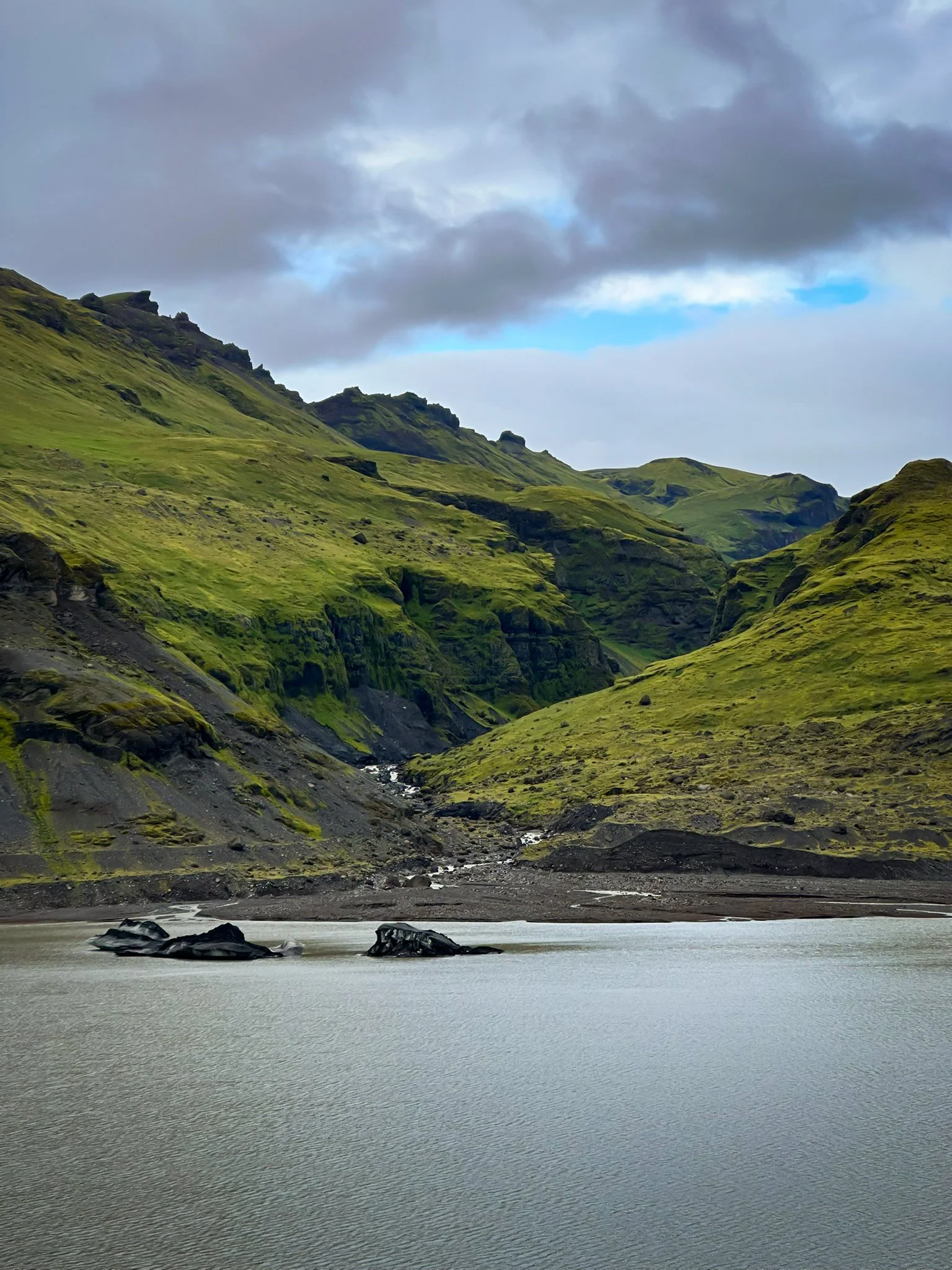 A scenic view of a lake with mountains covered in green grass and some rocks, under a cloudy sky with patches of blue.