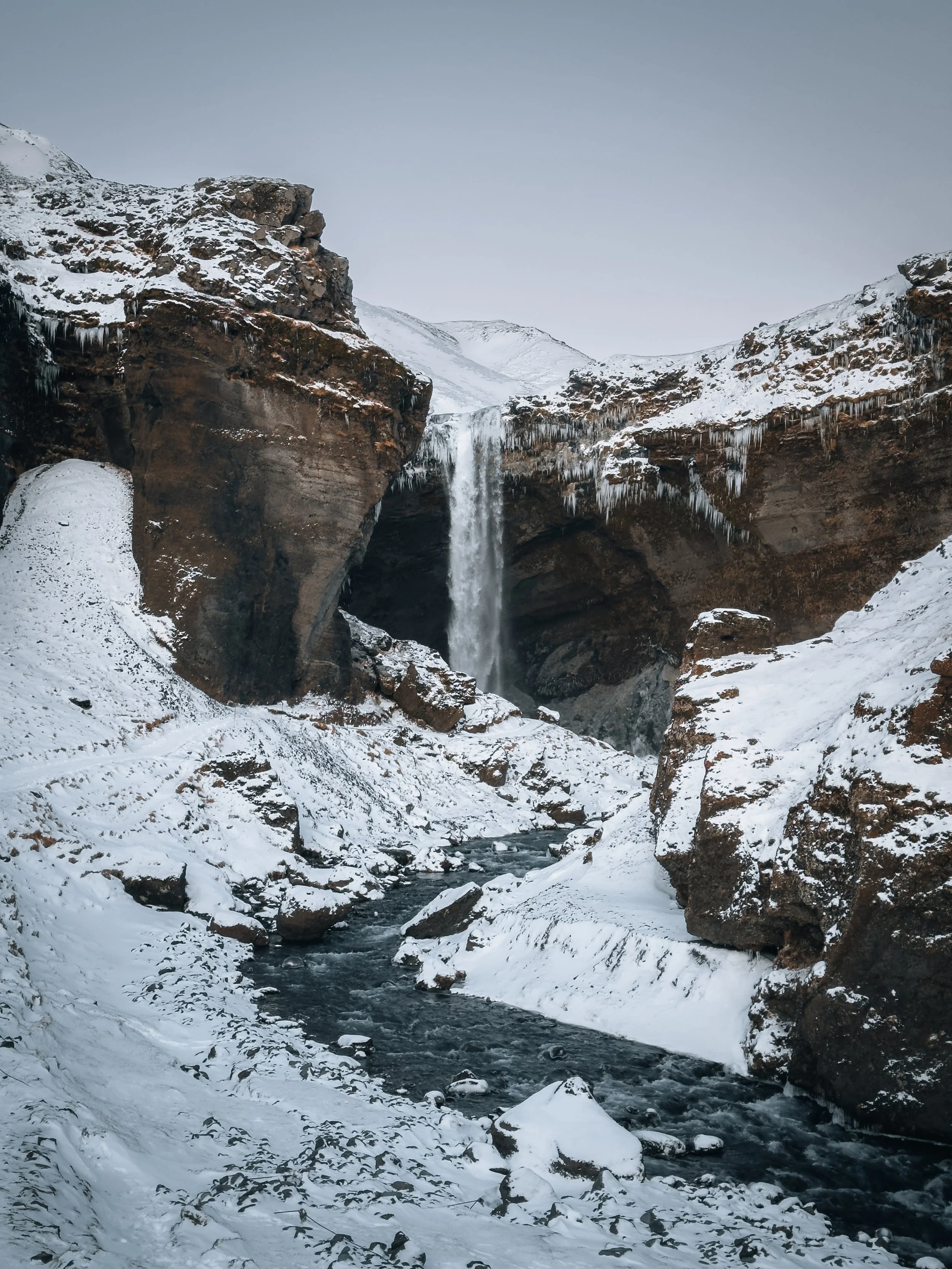 Snow-covered canyon with a waterfall flowing into a river, surrounded by icy rocks and cliffs under a cloudy sky.