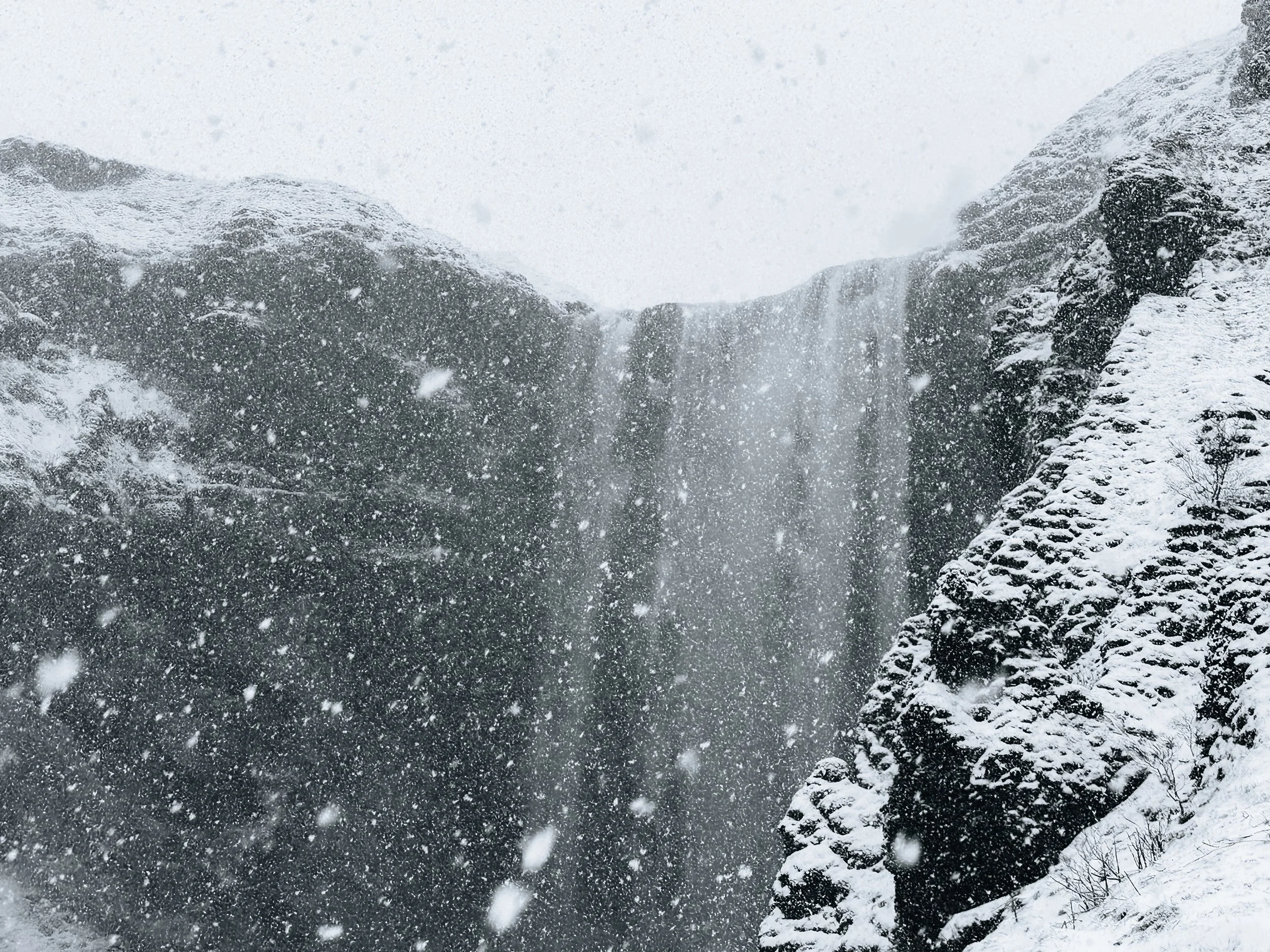 Snow falling at a waterfall surrounded by snow-covered rocks and cliffs.