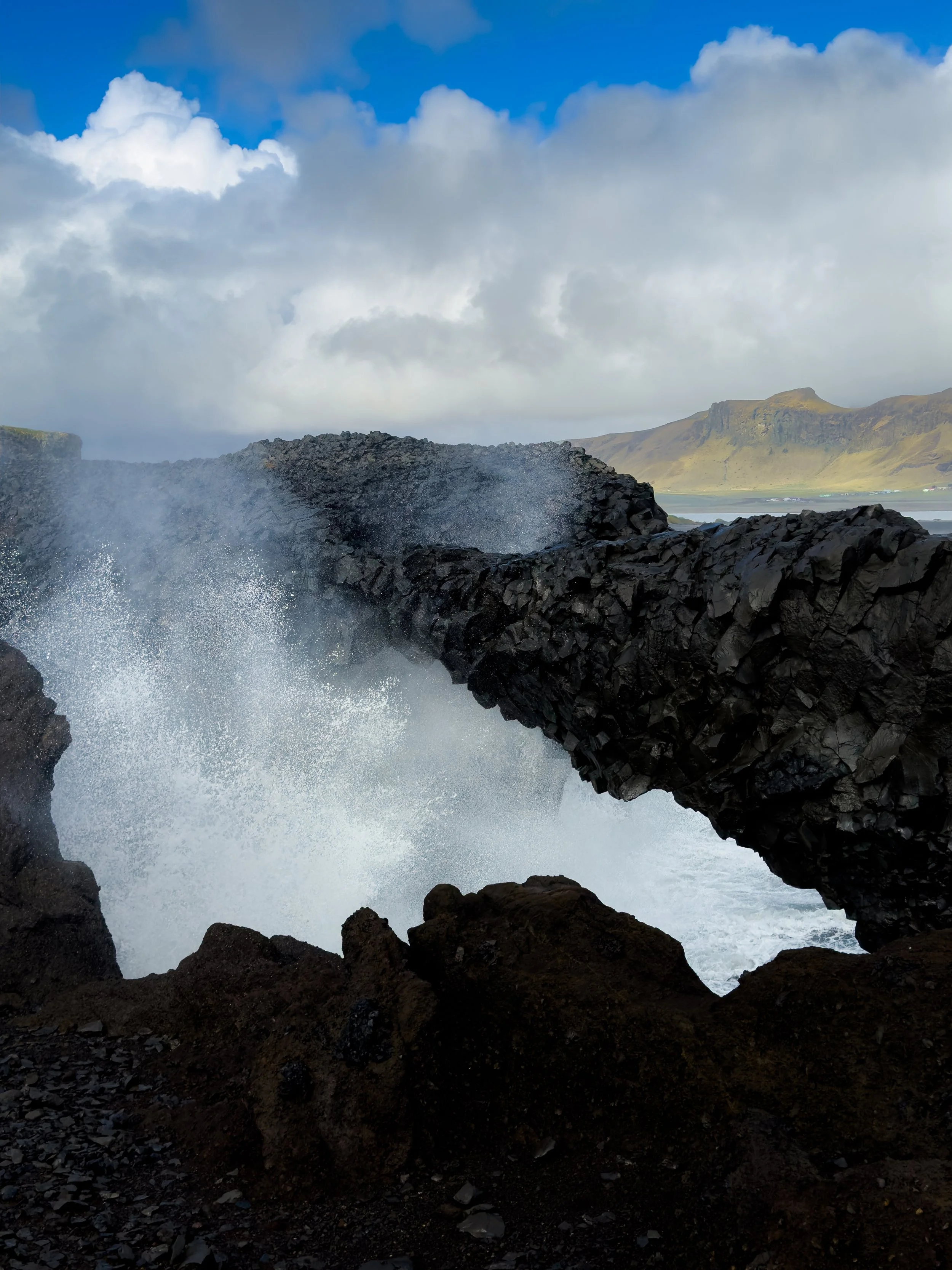 View of ocean waves crashing against rocky cliffs with a partly cloudy sky and distant mountains in the background.