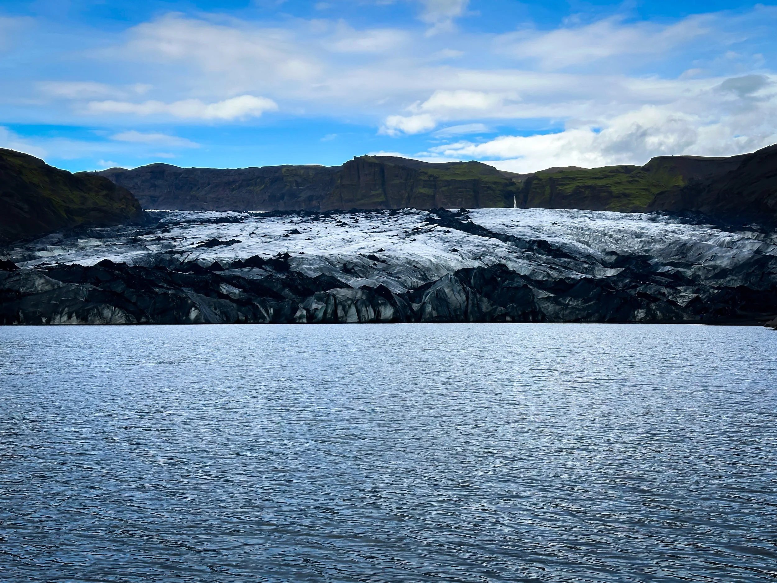 A body of water with a glacier in the background and grassy green hills on the sides under a partly cloudy sky.