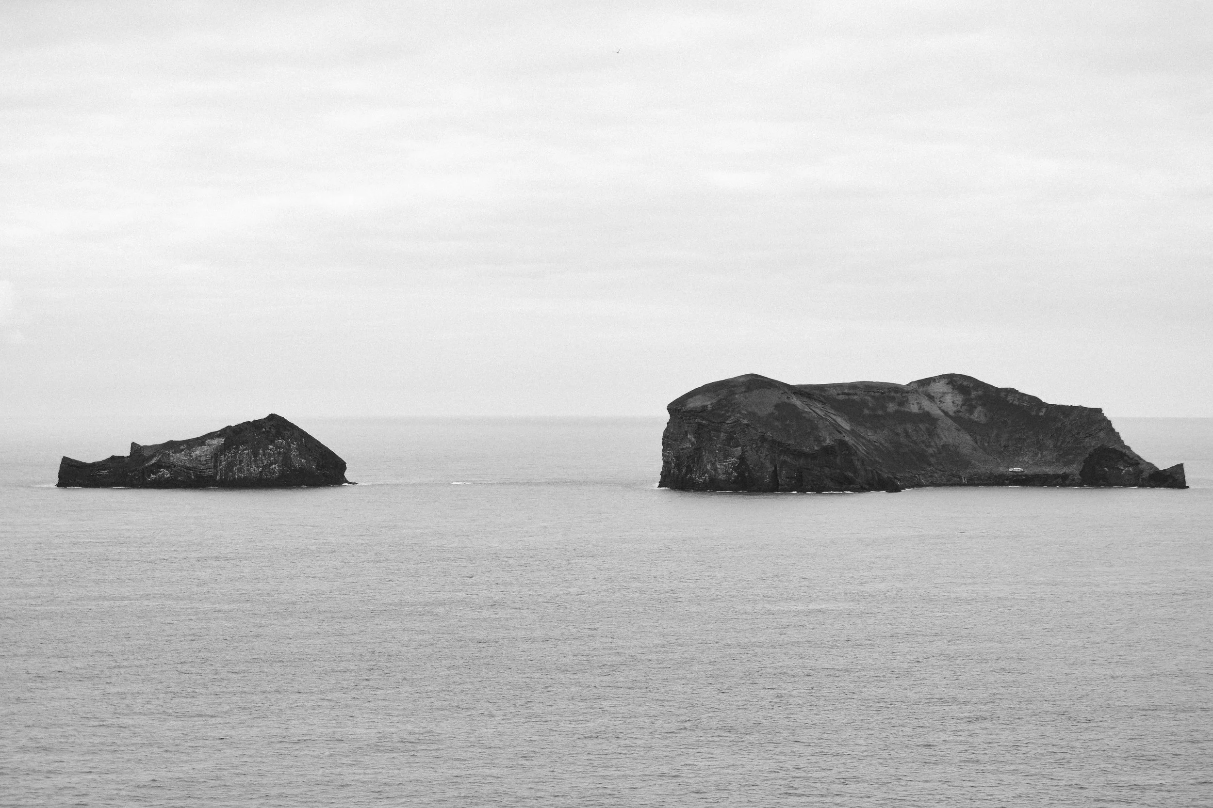 Black and white photo of two large rocky islands in the ocean, with a flat horizon and overcast sky.
