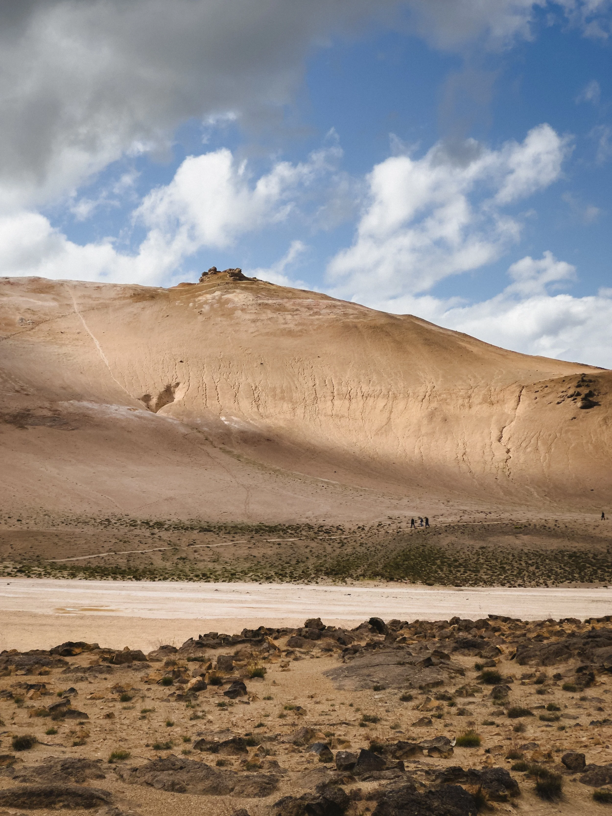 A desert landscape with a large sandy hill and a few hikers walking on a trail near the base. The sky is partly cloudy with blue patches.