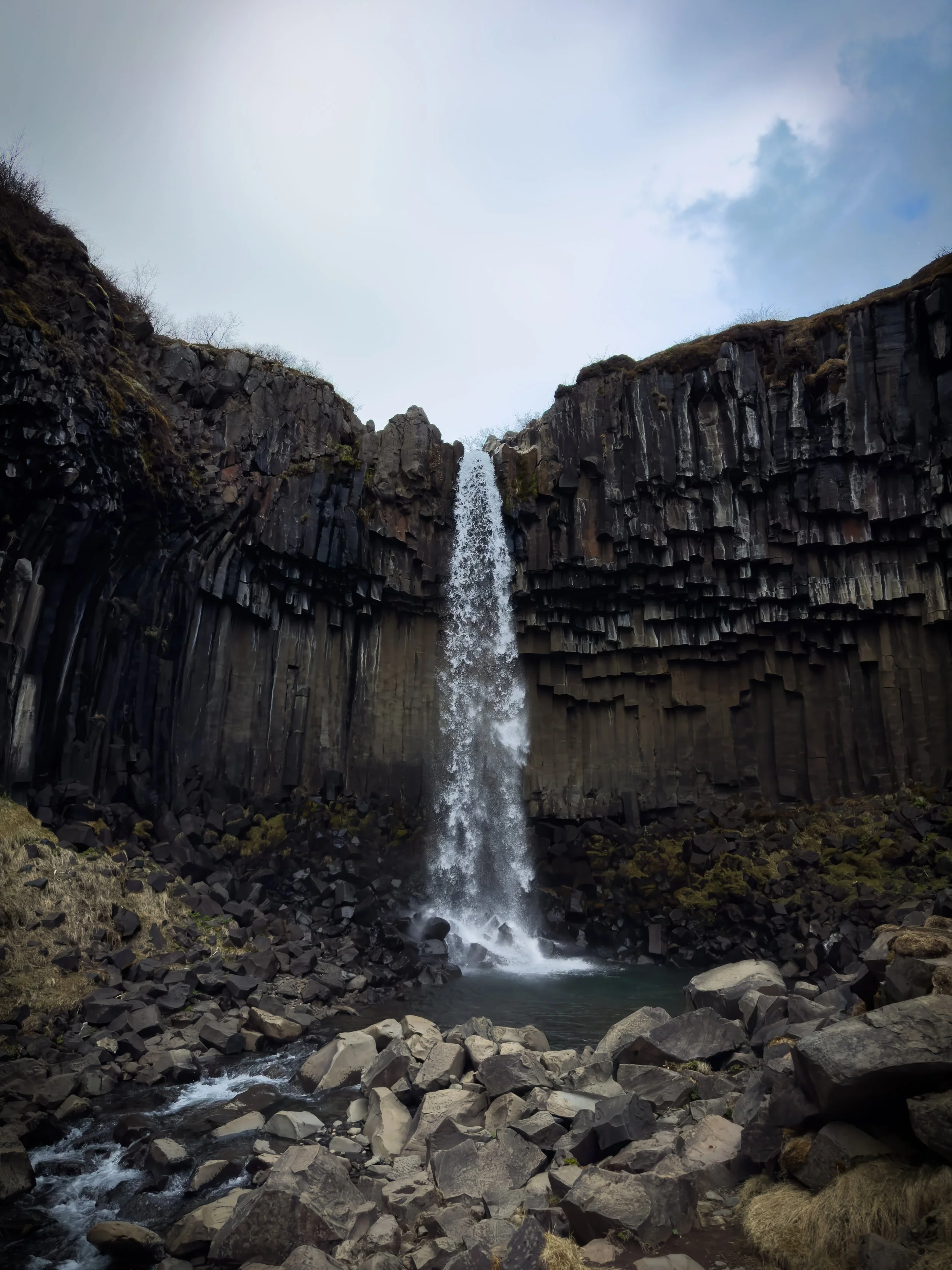A tall waterfall flowing into a small pool, surrounded by dark basalt columns and rocky terrain.