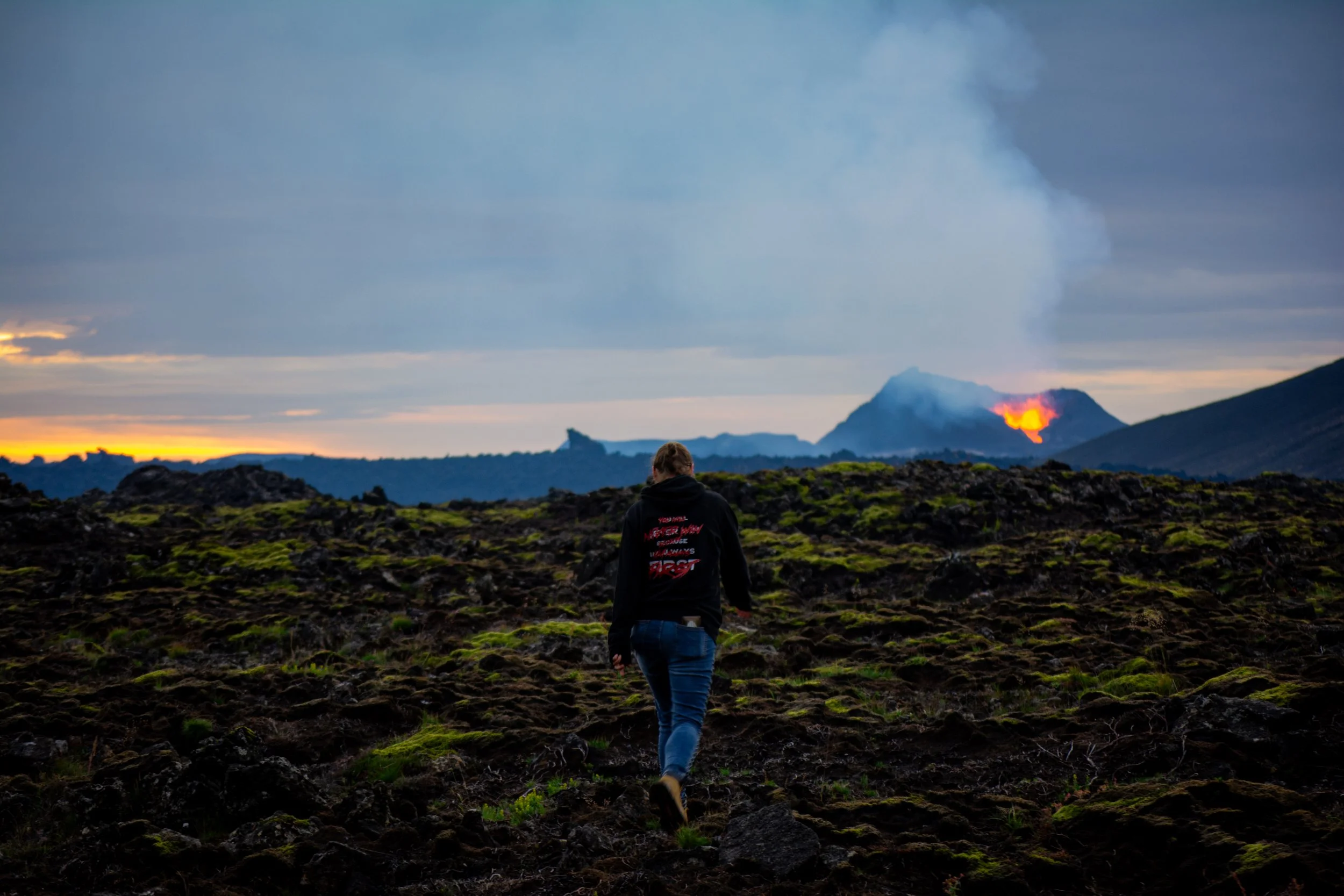 A person walking on a rocky landscape towards an erupting volcano with lava and smoke in the distance during sunset.