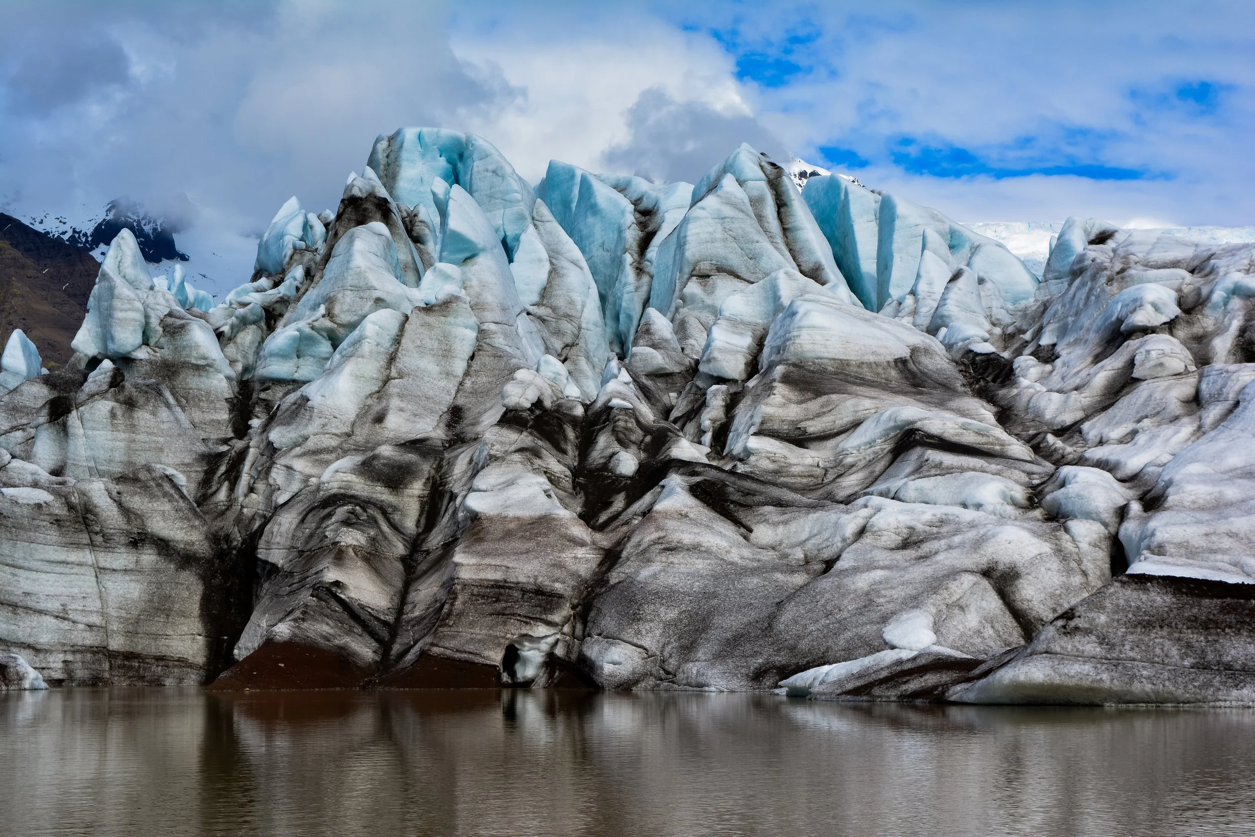 Glacier with ice formations and dark streaks near a body of water, cloudy sky in background.