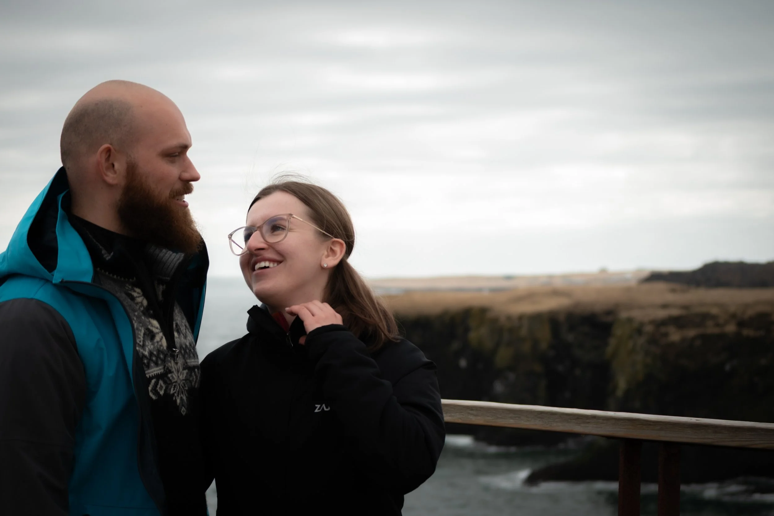 A man and woman are standing close together outdoors on a cloudy day, smiling and looking at each other, with a landscape of water and land in the background.