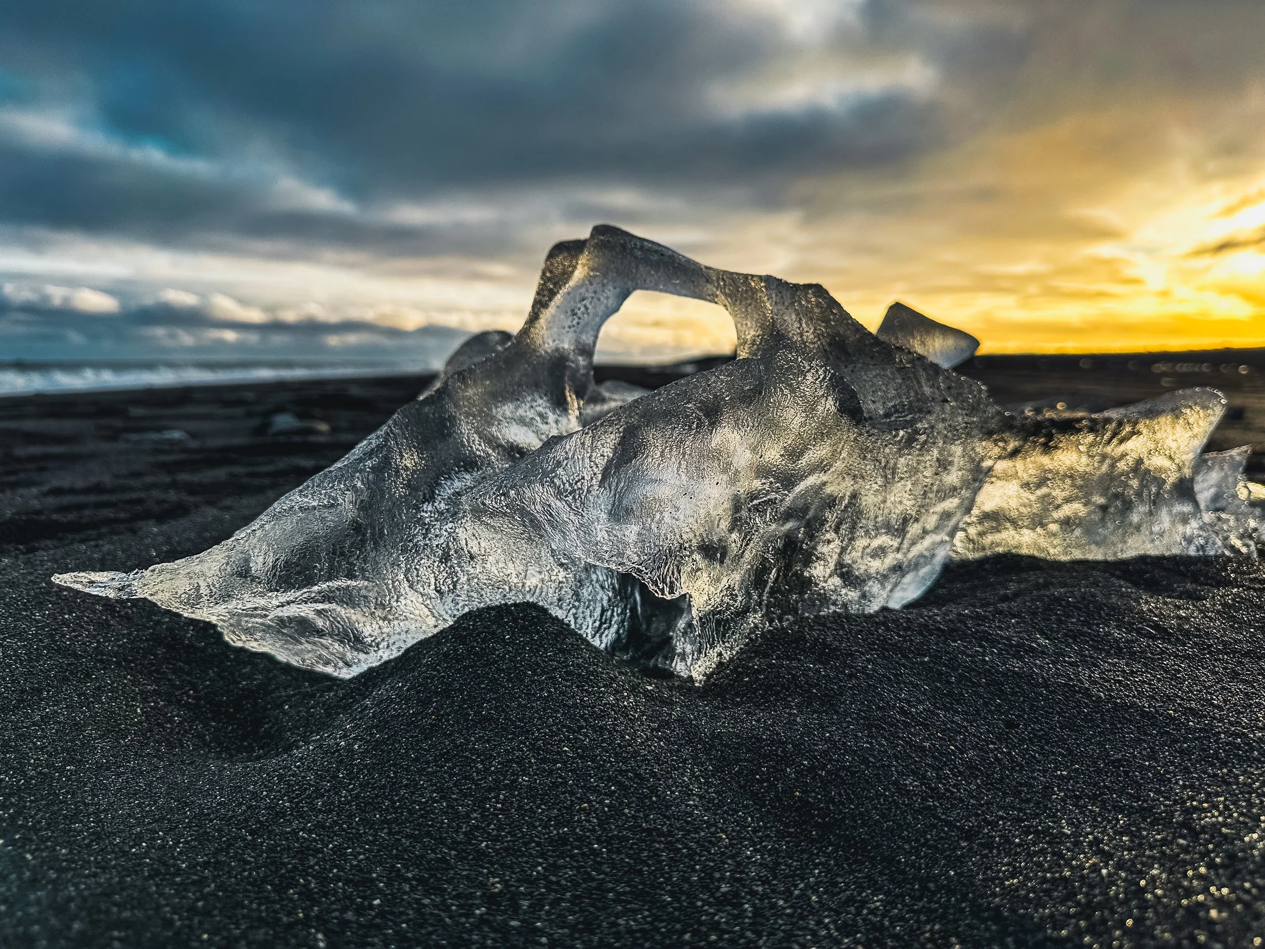 A large, clear ice fragment on black sand beach during sunset with cloudy sky.