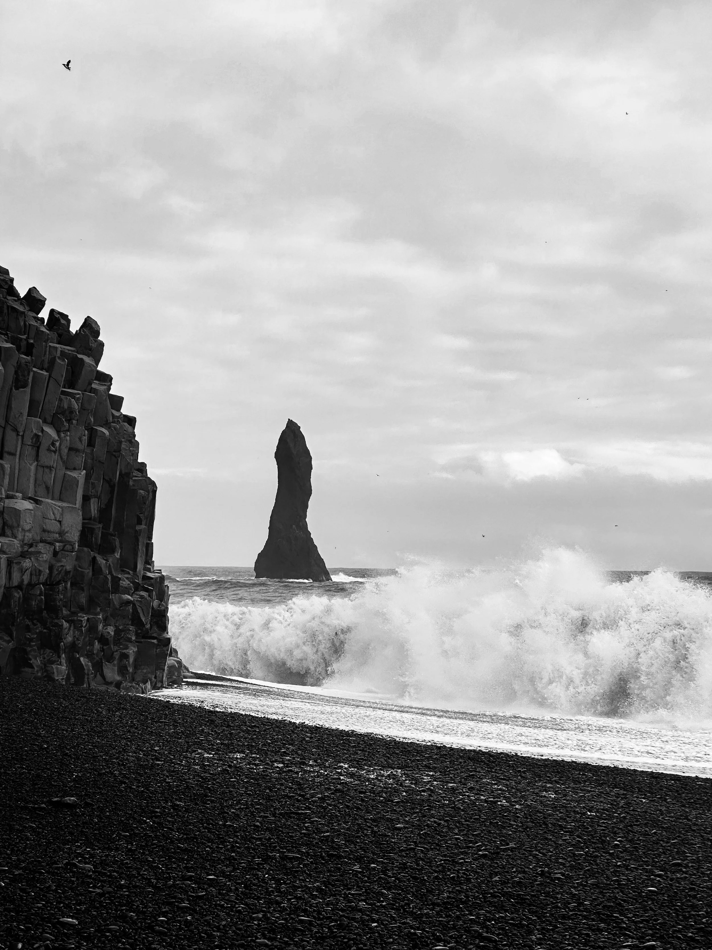 Black-and-white photo of a rocky cliff on the left, a large rock formation in the ocean in the distance, and waves crashing onto a dark, pebbly beach under a cloudy sky.