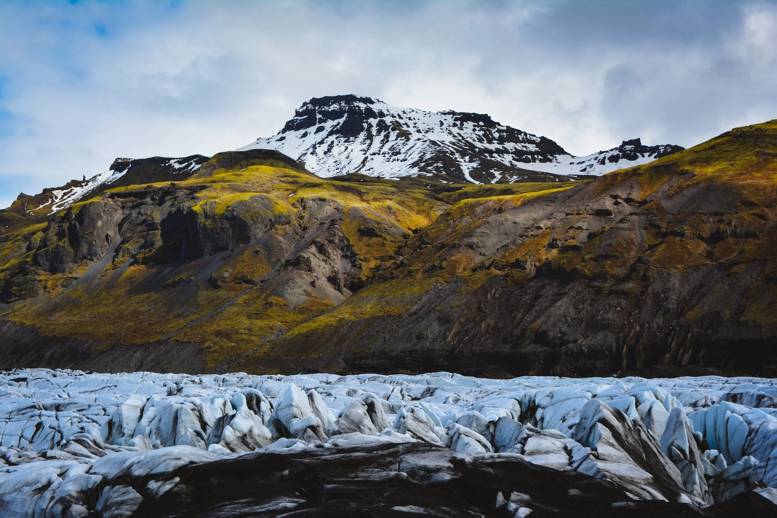 A landscape of a mountain with snow patches, a cloudy sky, and a foreground of a glacier with blue and white ice formations.
