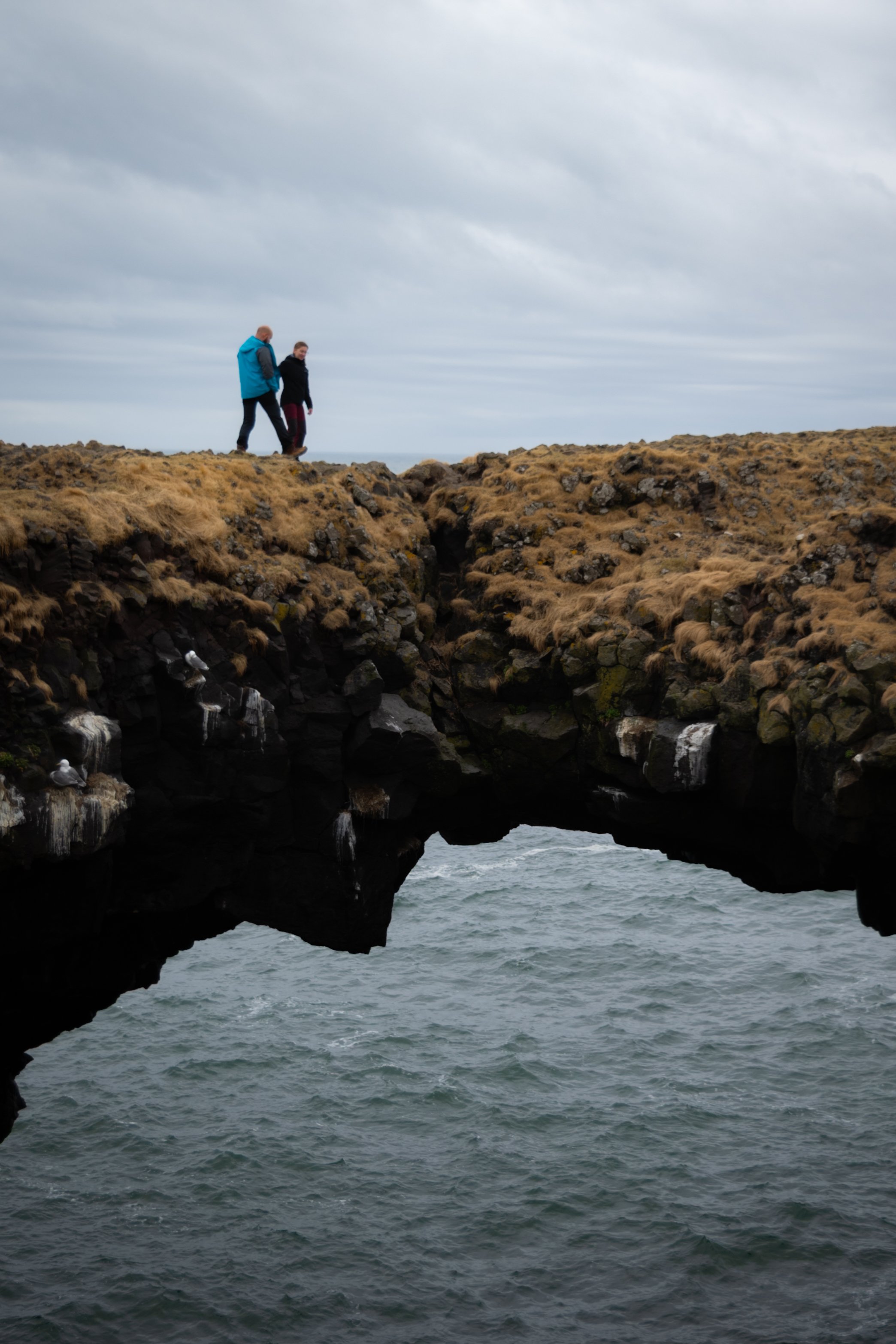 Two people walk along a rocky, grassy coastal area with an archway formed by rocks above the ocean, under a cloudy sky.