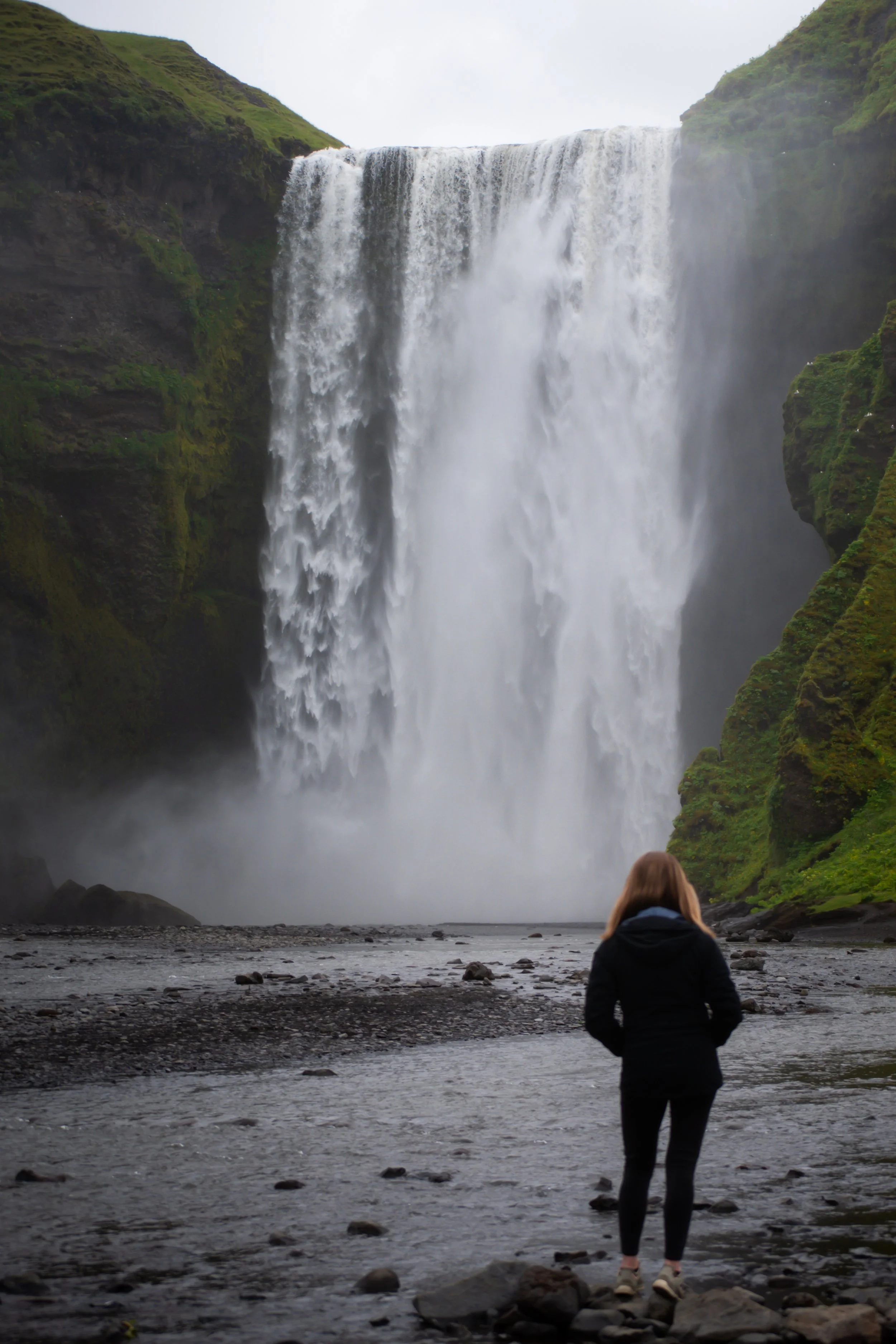 Person with brown hair and a dark jacket standing on rocks and looking at a large waterfall with flowing water and green moss-covered cliffs around it.