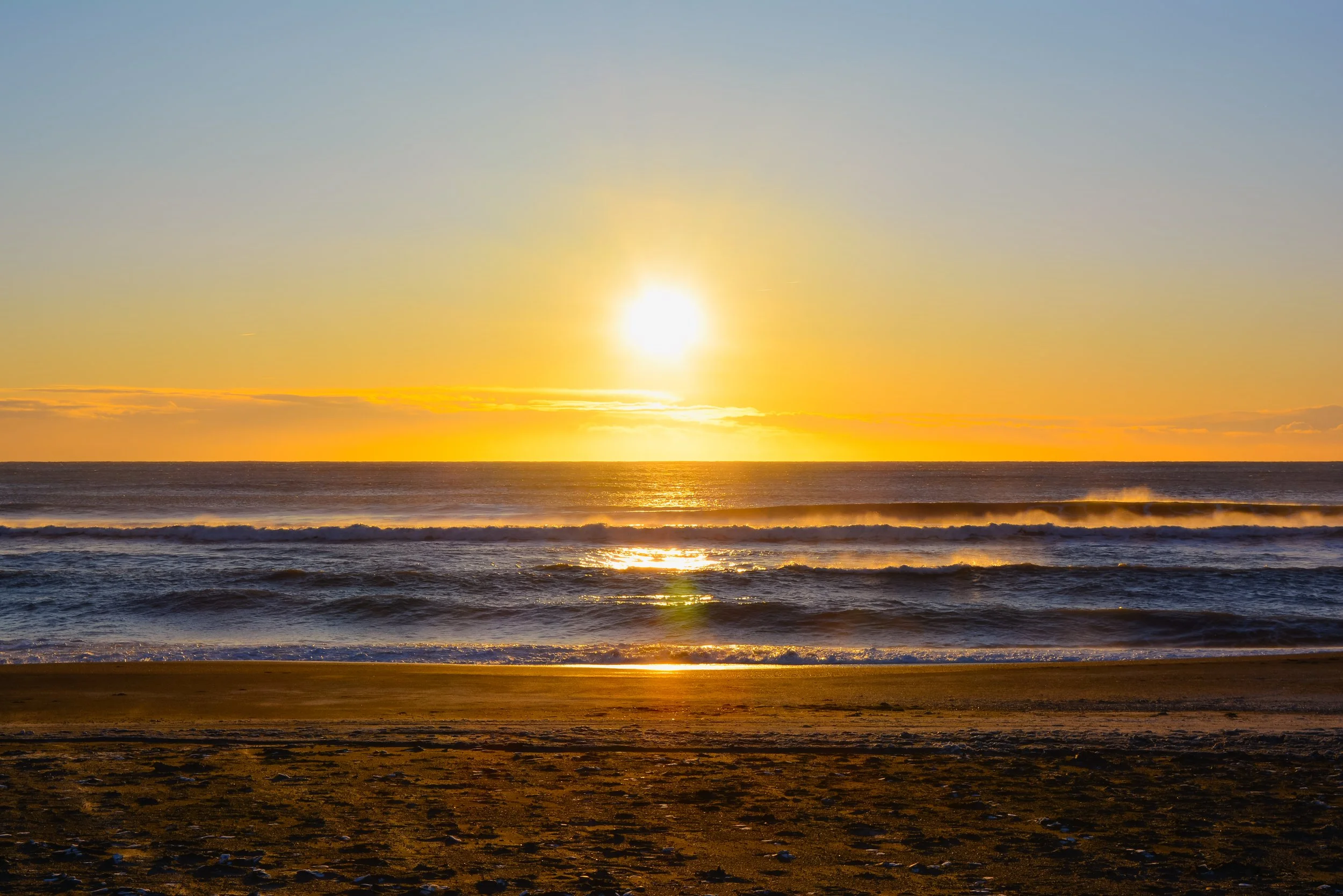 Sunset over the ocean with waves and a sandy beach in the foreground.