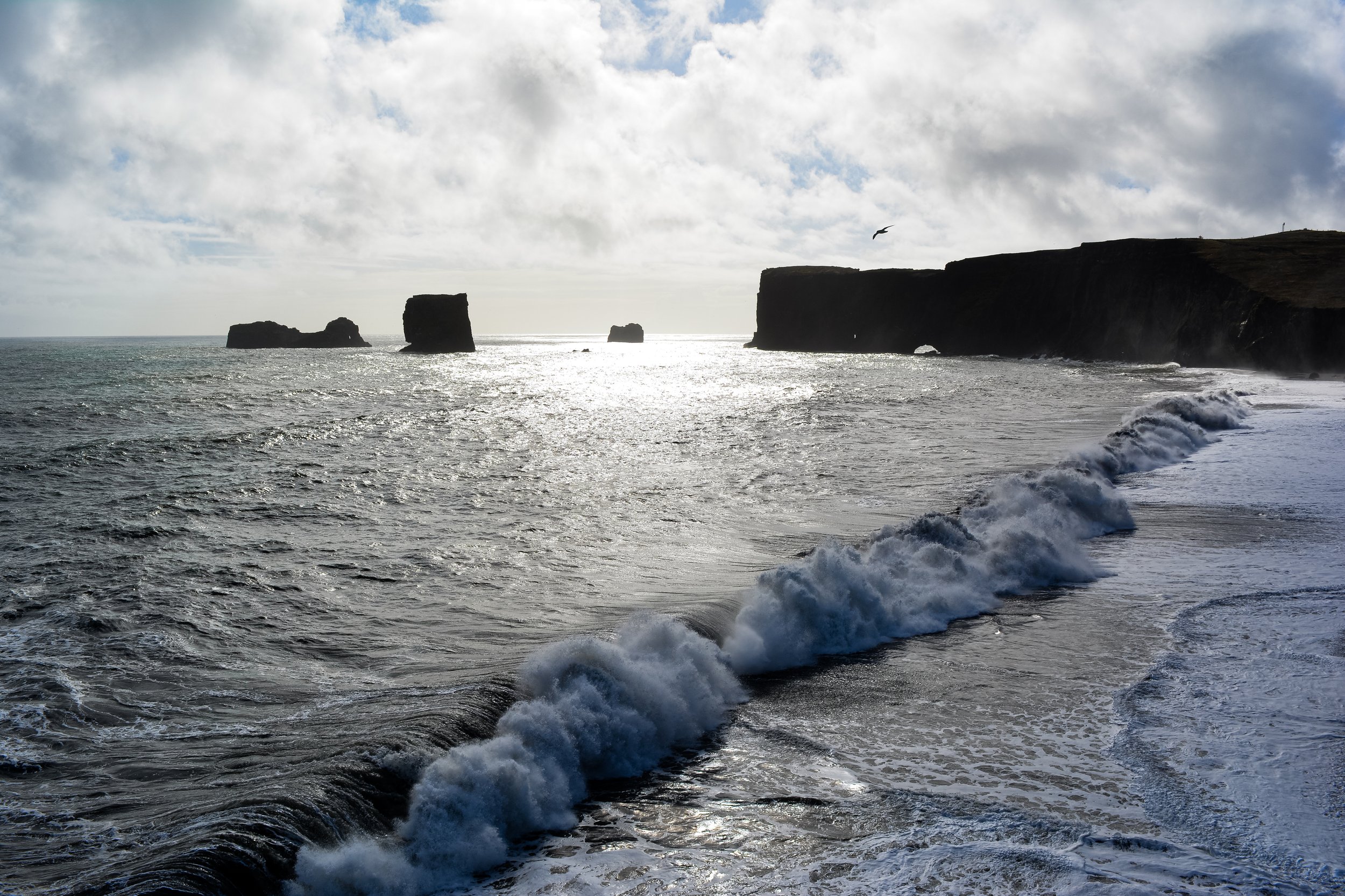 Ocean waves crashing in front of sea stacks and cliffs under a cloudy sky with sunlight breaking through.