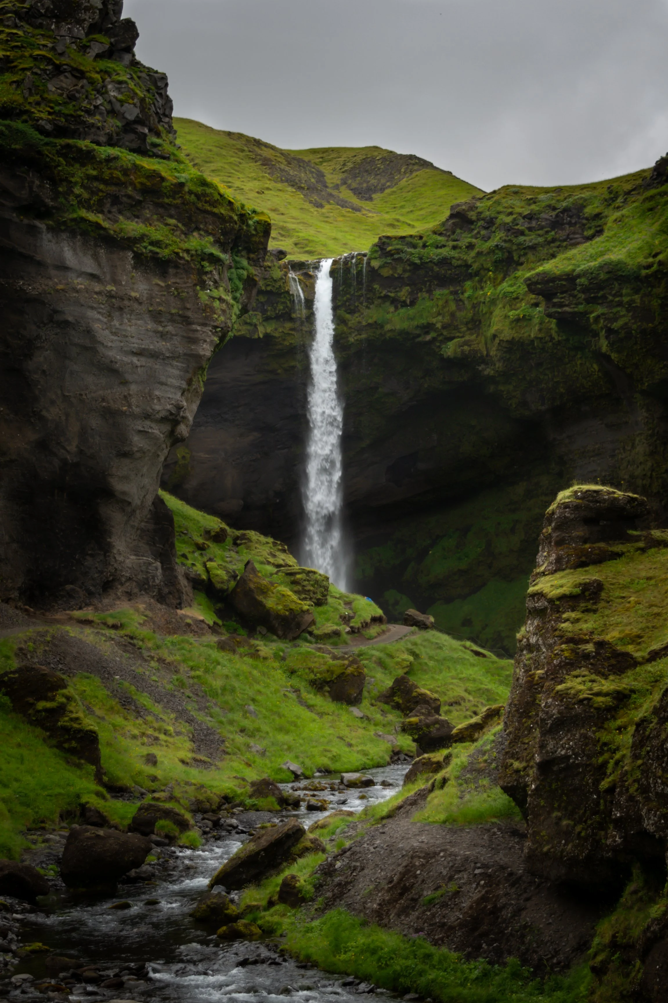 A scenic view of a tall waterfall cascading down a rocky cliffside in a lush green valley with a stream flowing through moss-covered rocks.