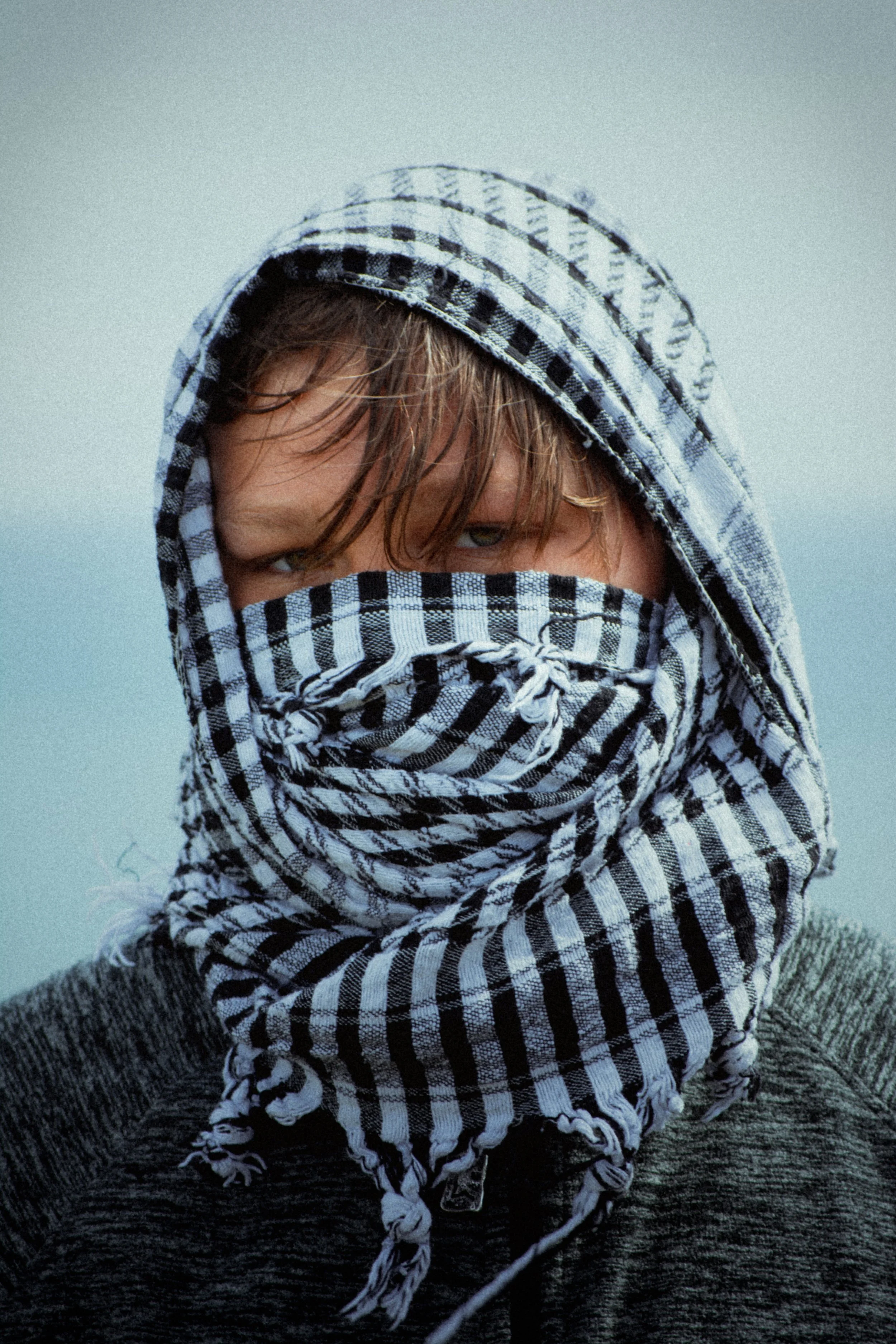 Person with wet hair wearing a black and white checkered scarf over face and head, gray sweater, against a cloudy sky.