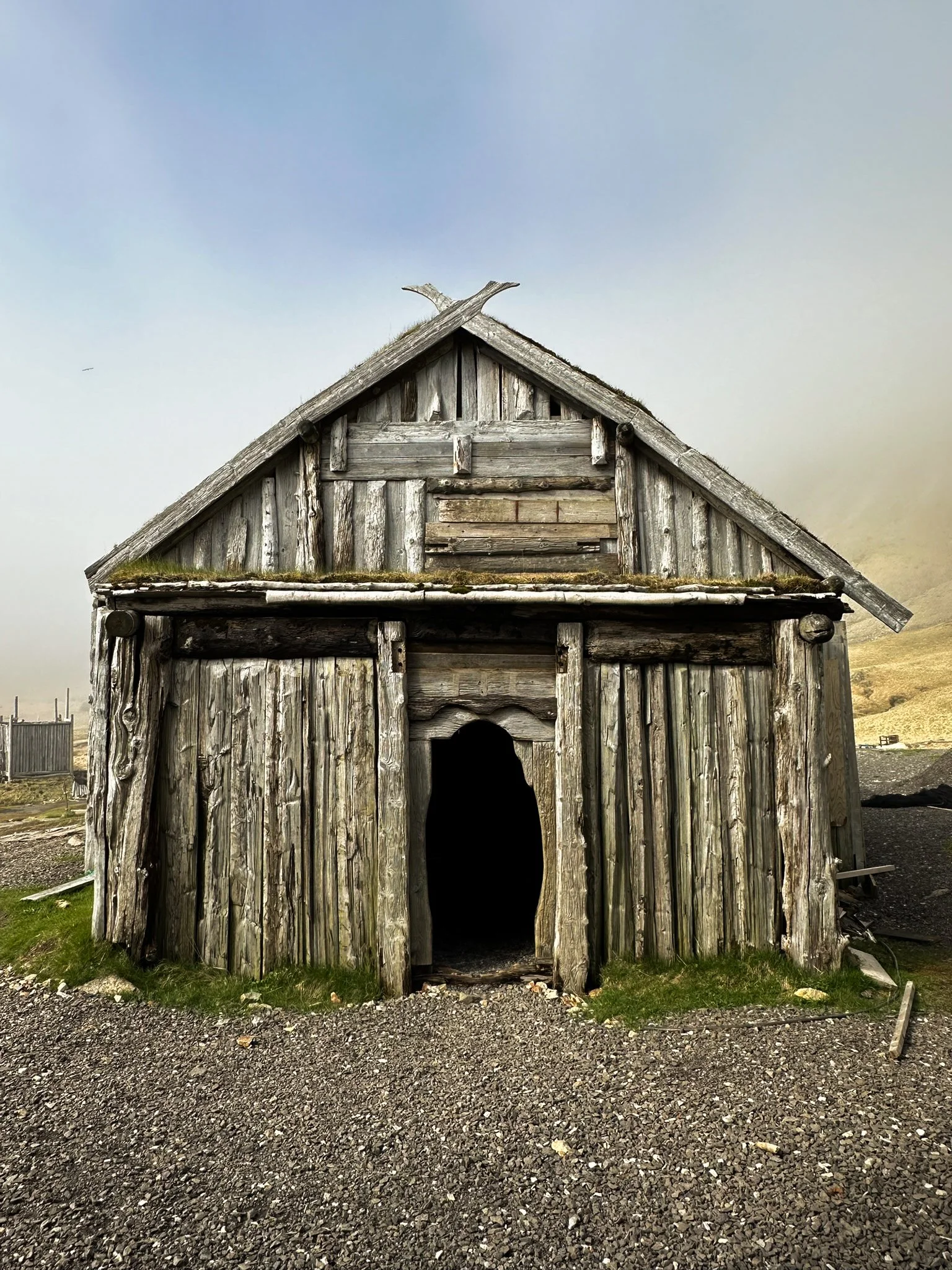 Old, weathered wooden cabin with a peaked roof, situated on a gravel ground with patches of grass. The cabin appears abandoned and has a dark open doorway.