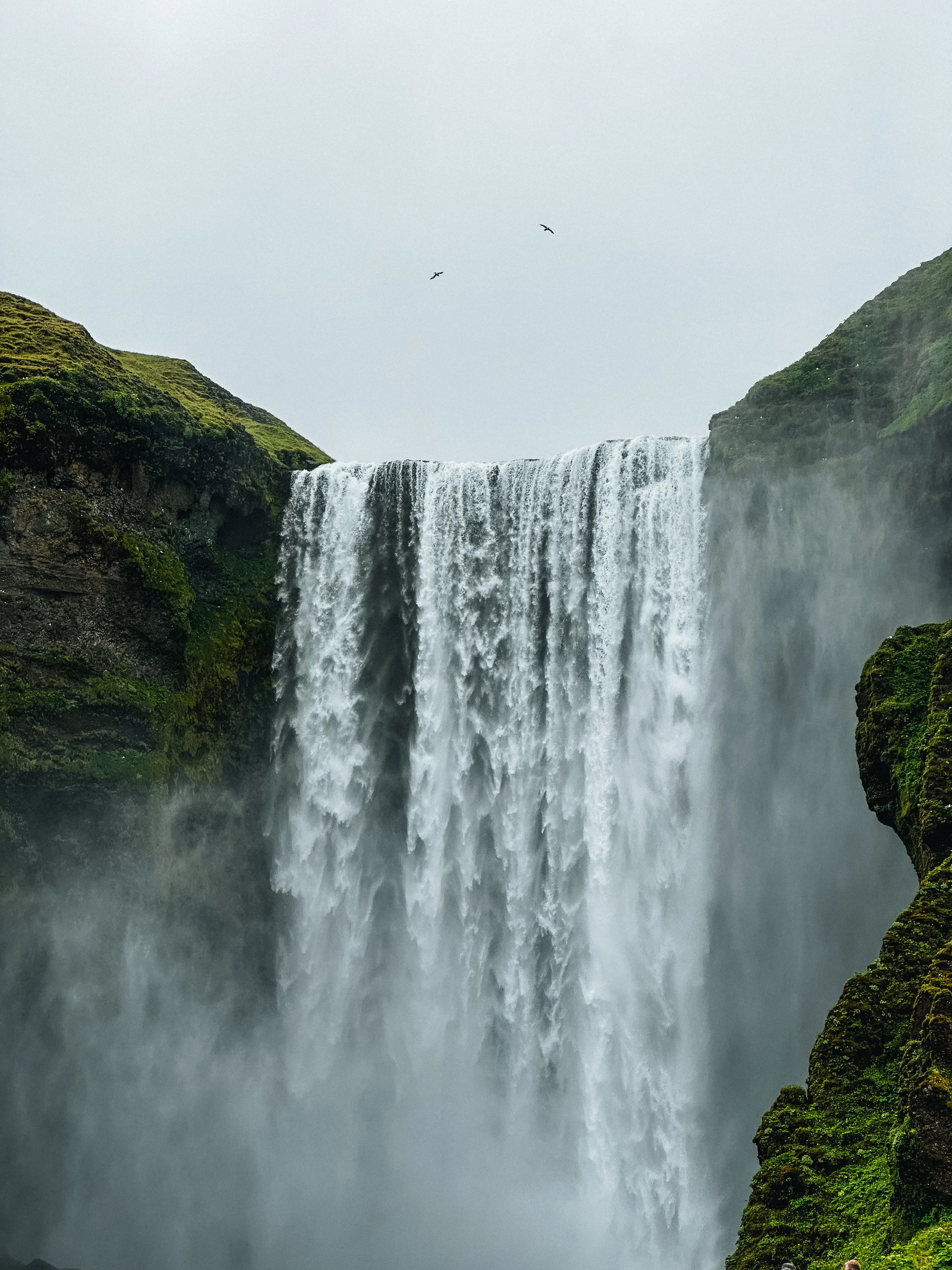 Waterfall flowing over moss-covered rocks with birds flying overhead in a cloudy sky.