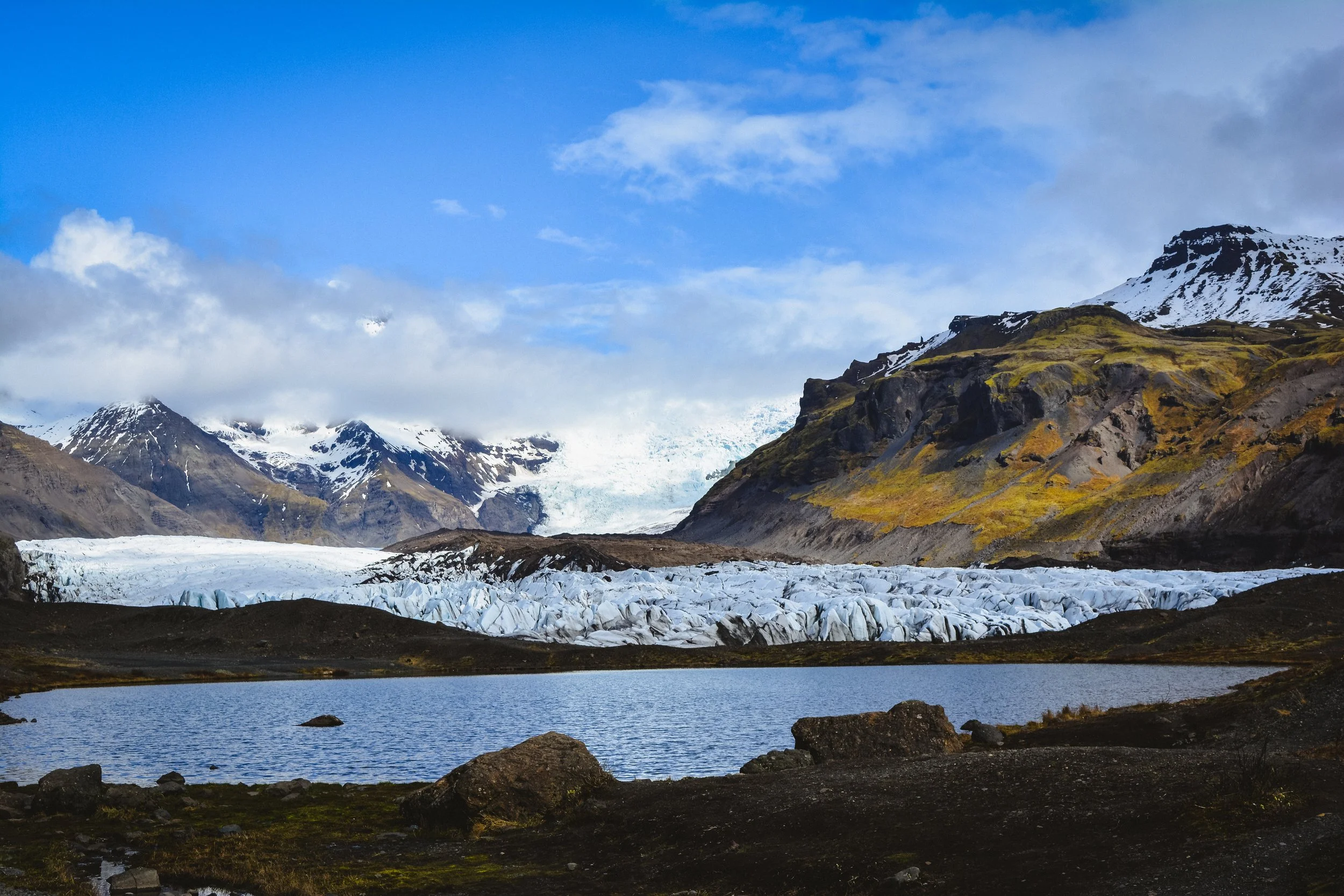 A scenic landscape of mountains with snow-capped peaks, a glacier, a lake in the foreground, and a partly cloudy sky.