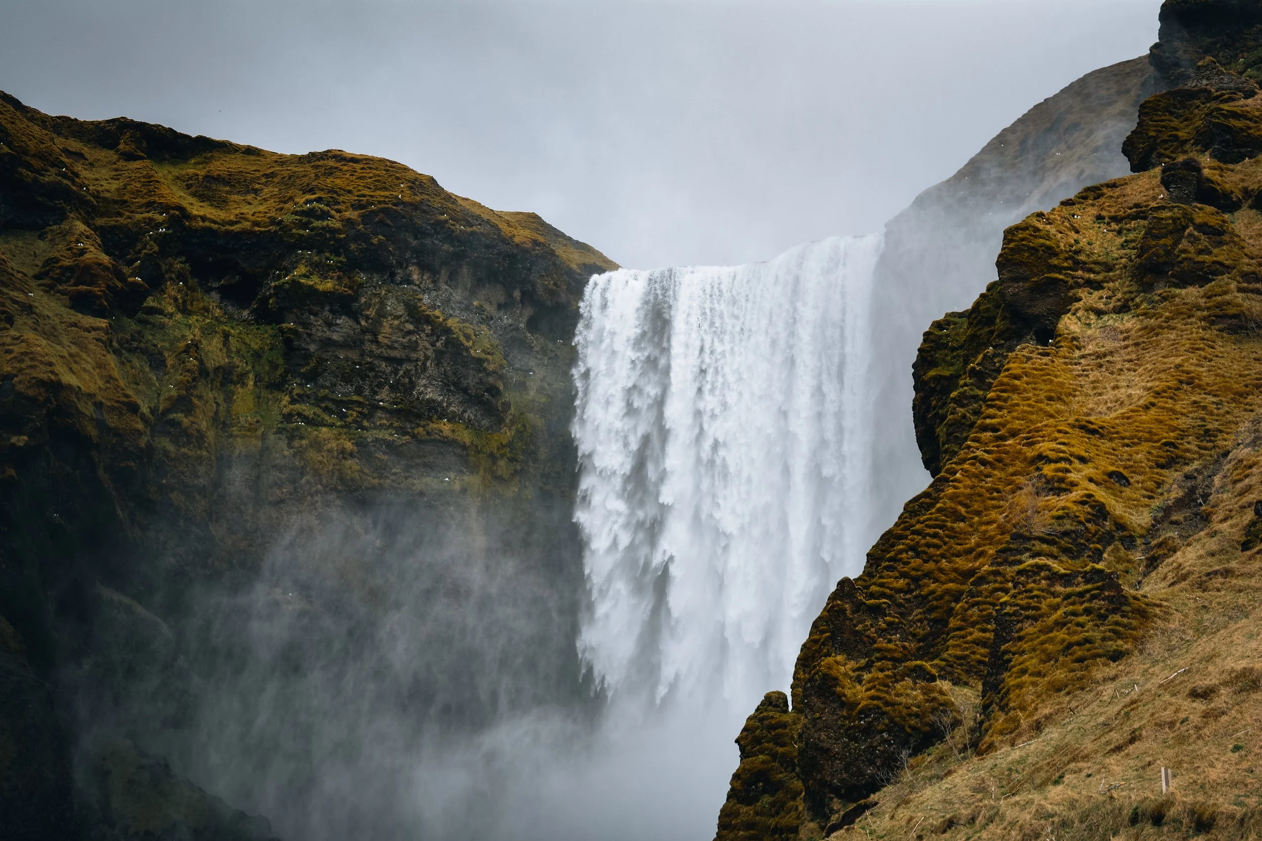 A large waterfall flowing over a cliff into a misty pool below, surrounded by moss-covered rocky cliffs and grassy landscape under an overcast sky.
