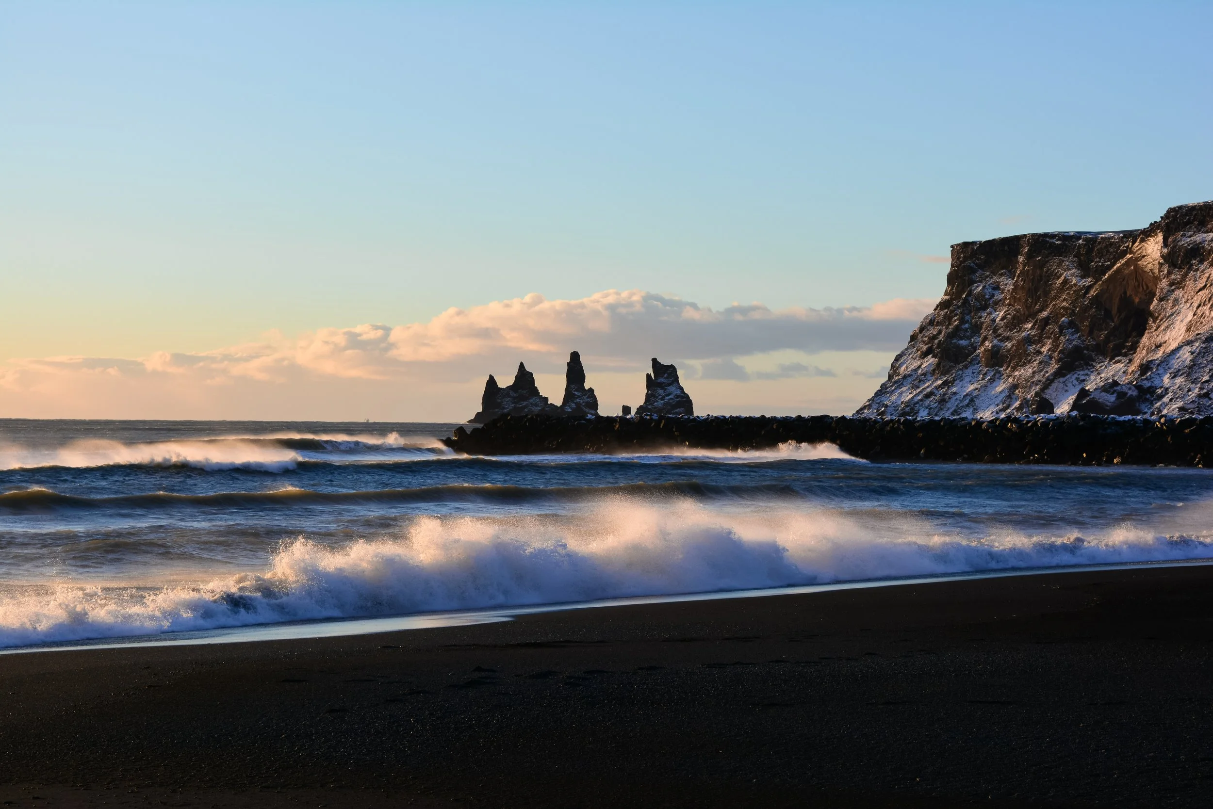 Black sand beach with waves crashing against the shore and rock formations in the water, with a large rocky cliff on the right side, during sunset or sunrise.