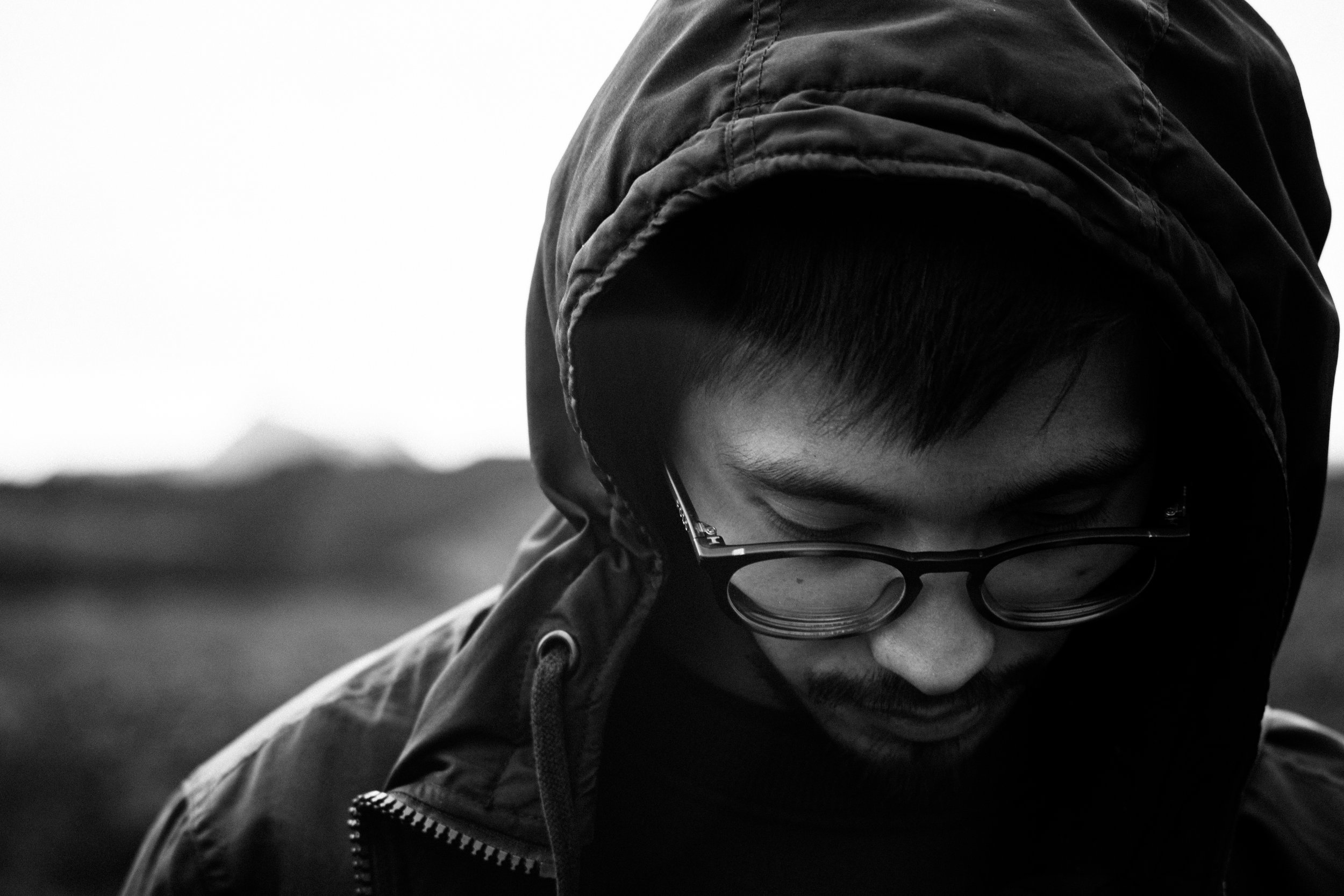 Black and white close-up photo of a man with glasses, hooded jacket, and dark hair, looking downward outdoors.