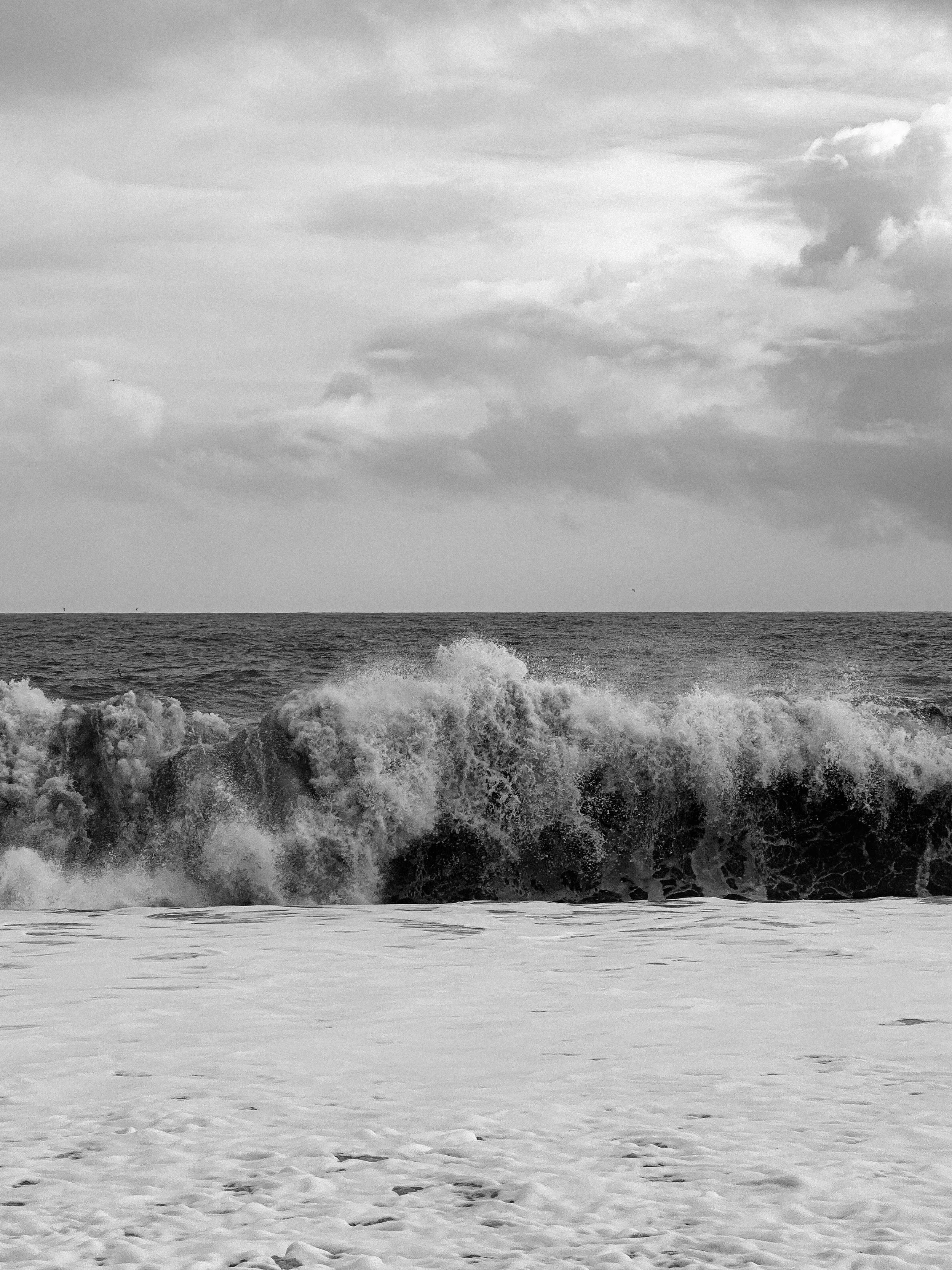 Black and white photo of ocean waves crashing onto the sandy shore with cloudy sky overhead.
