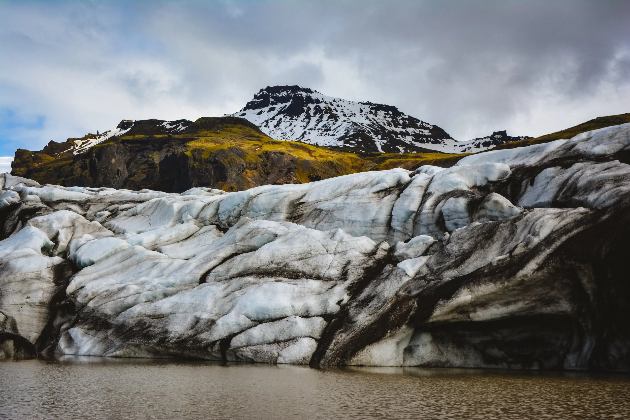 A glacier with snow and ice, flowing into a body of water, with a mountain in the background partly covered in snow and clouds overhead.