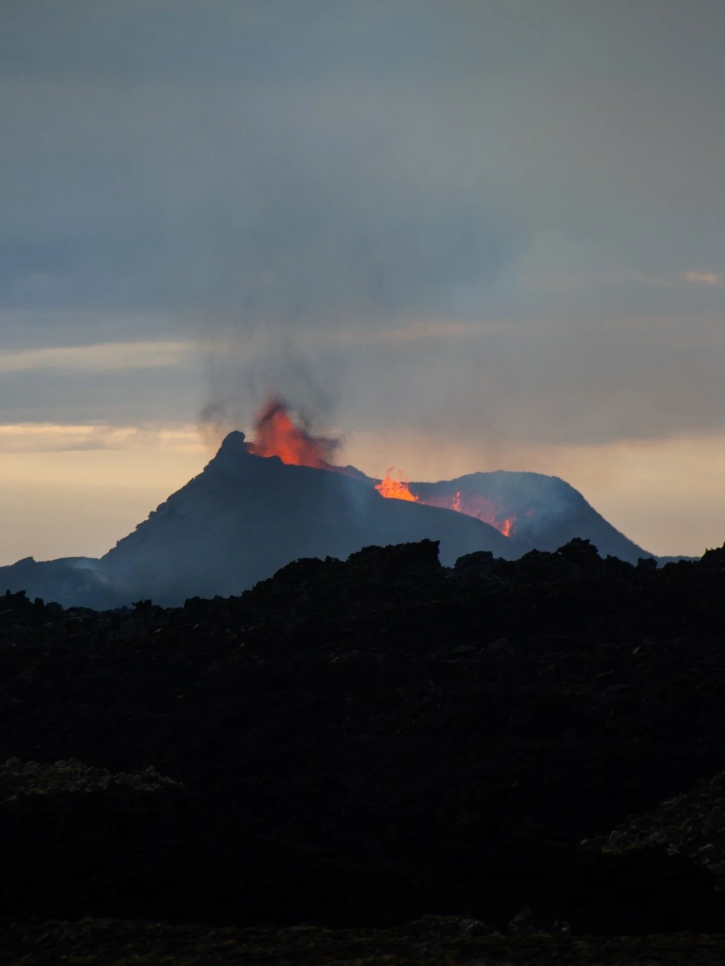 Active volcano erupting with lava and smoke, dark rocky terrain in the foreground, cloudy sky.