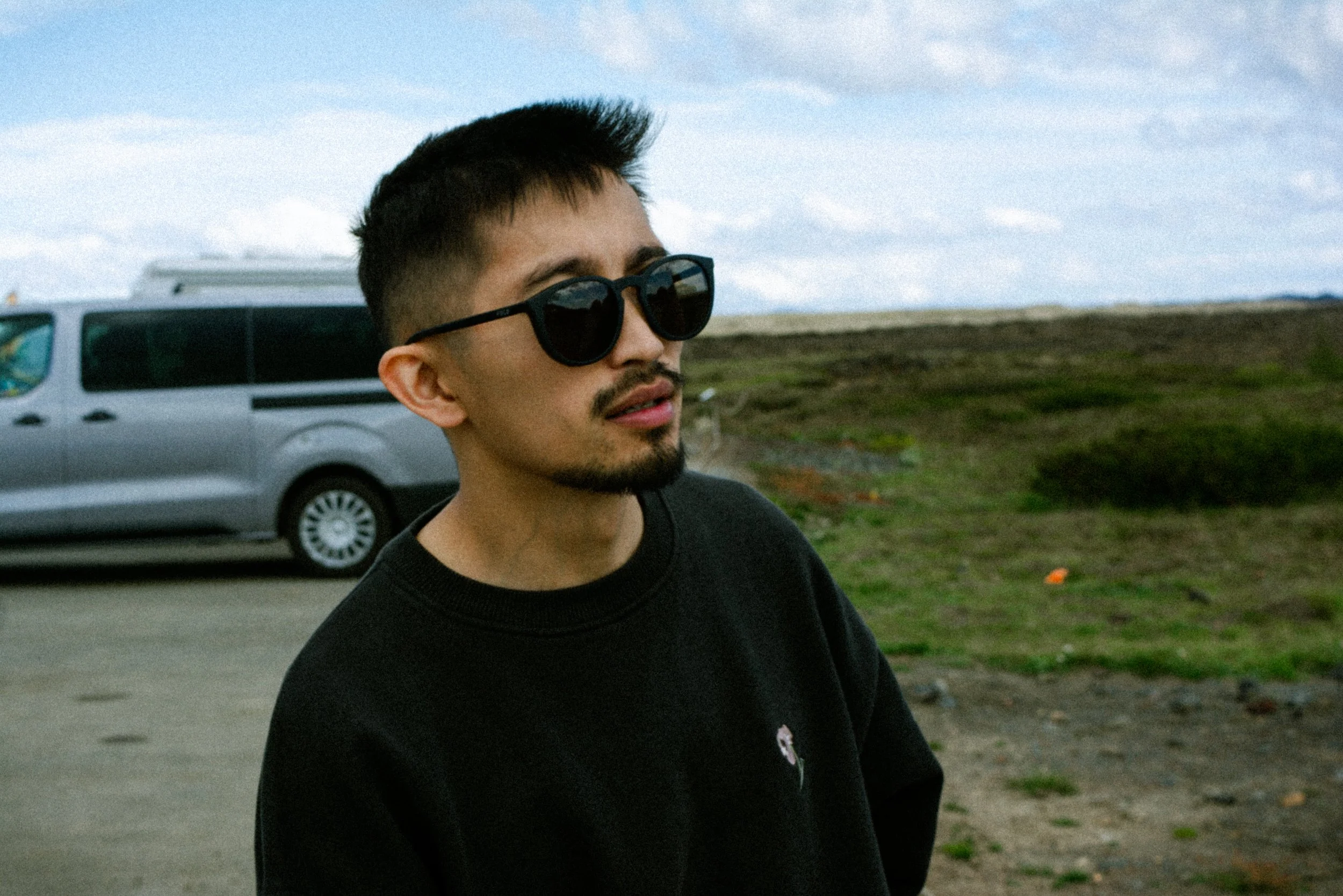Young man with short hair, sunglasses, and a goatee, standing outdoors in front of a van and open landscape.