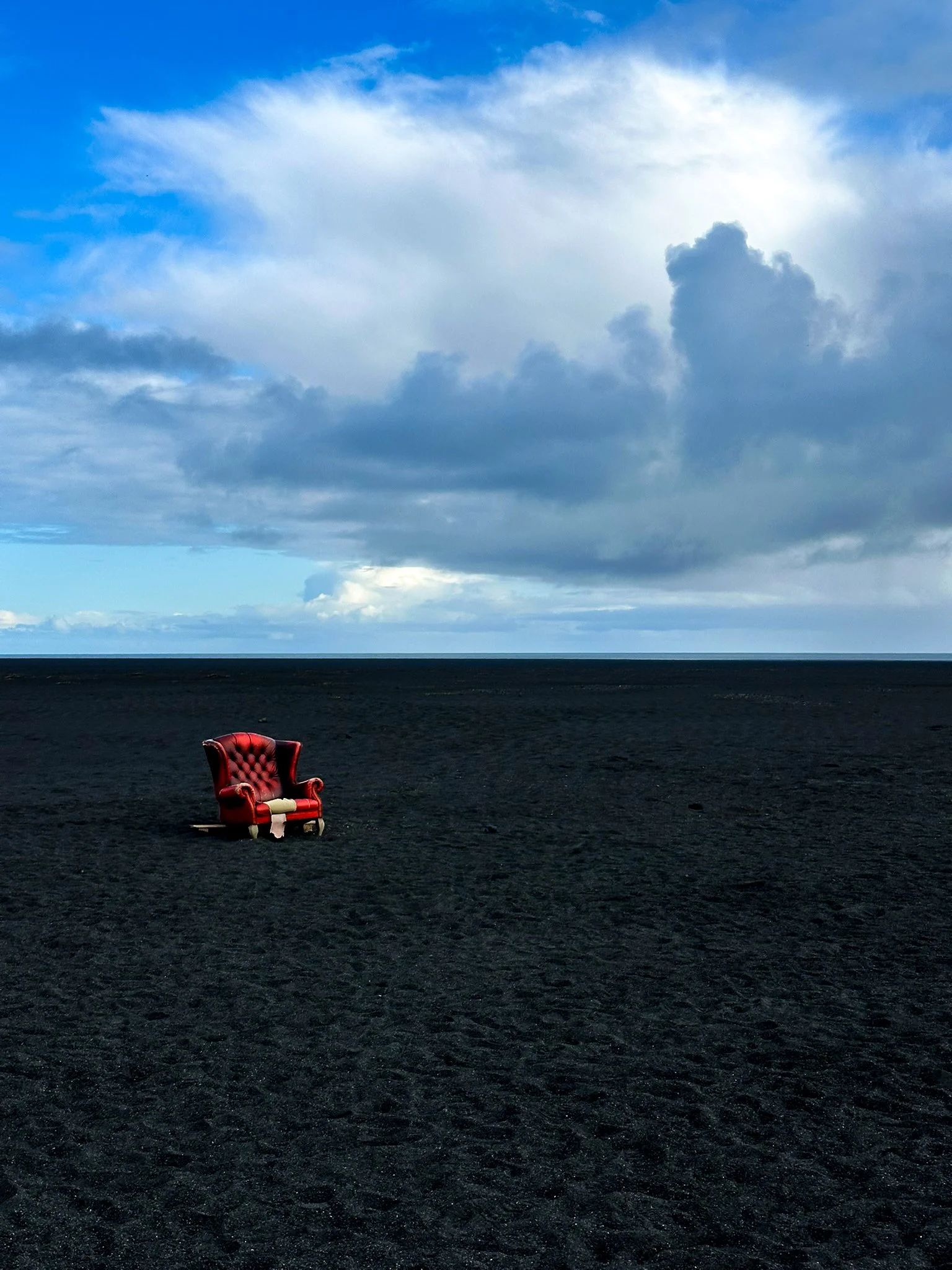A red armchair on a dark, almost black, sandy beach under a cloudy sky.