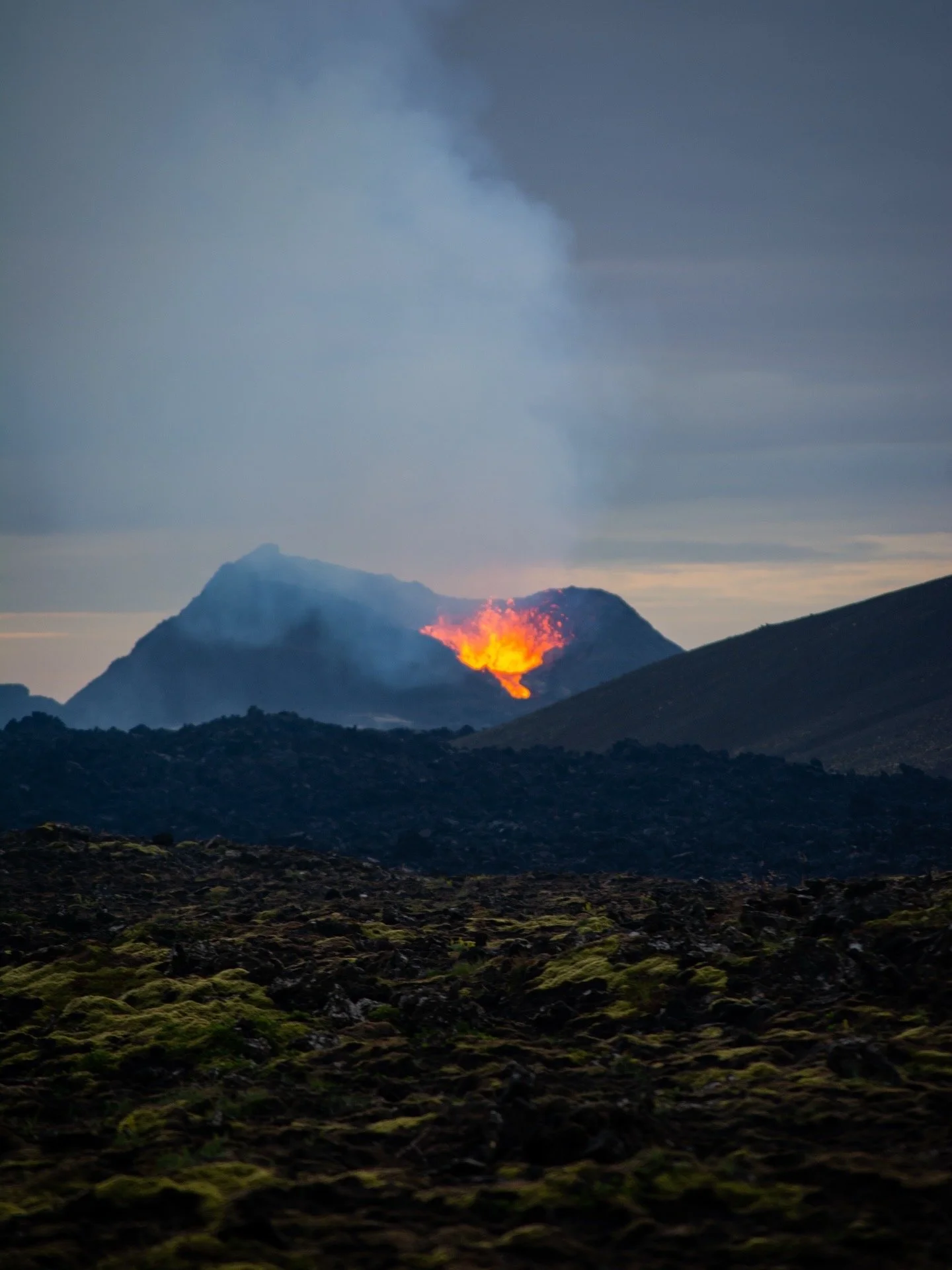 Active volcano erupting with lava flowing and smoke rising in the background, dark landscape in the foreground.
