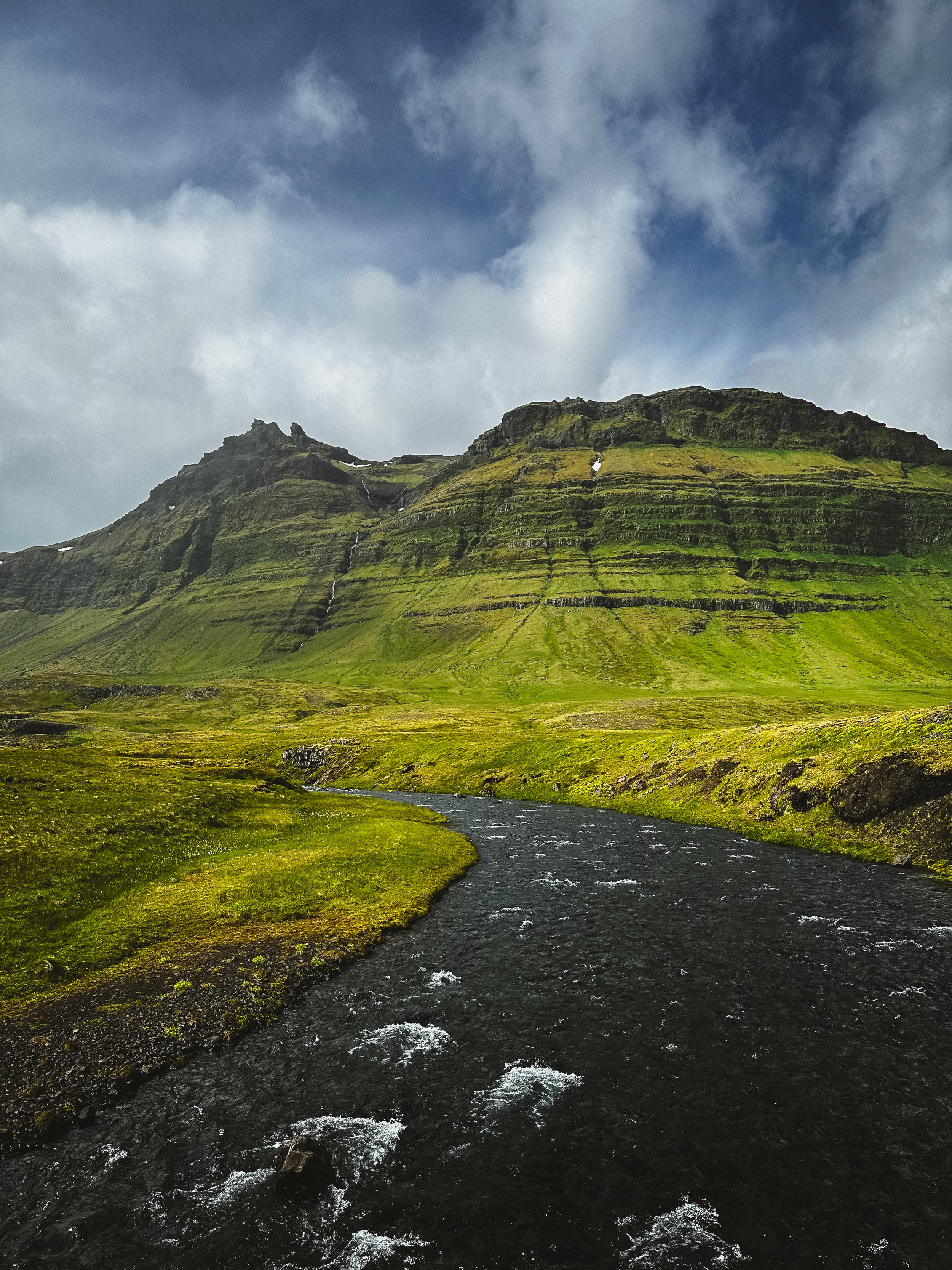 A scenic landscape features a river flowing through green grassy fields towards a mountain with layered slopes and patches of snow, under a cloudy sky.