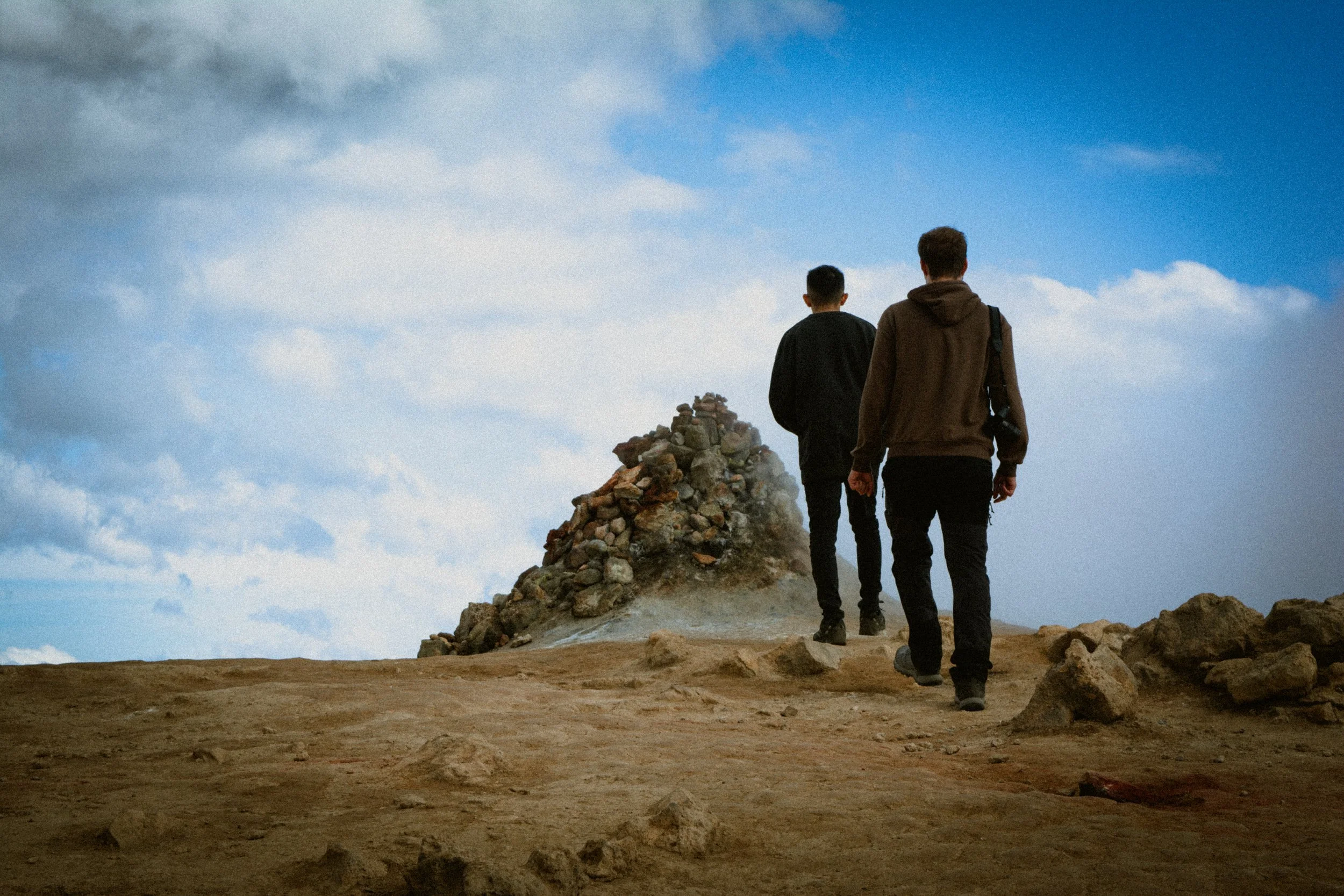 Two people walking towards a rocky summit on a barren landscape under a partly cloudy sky.