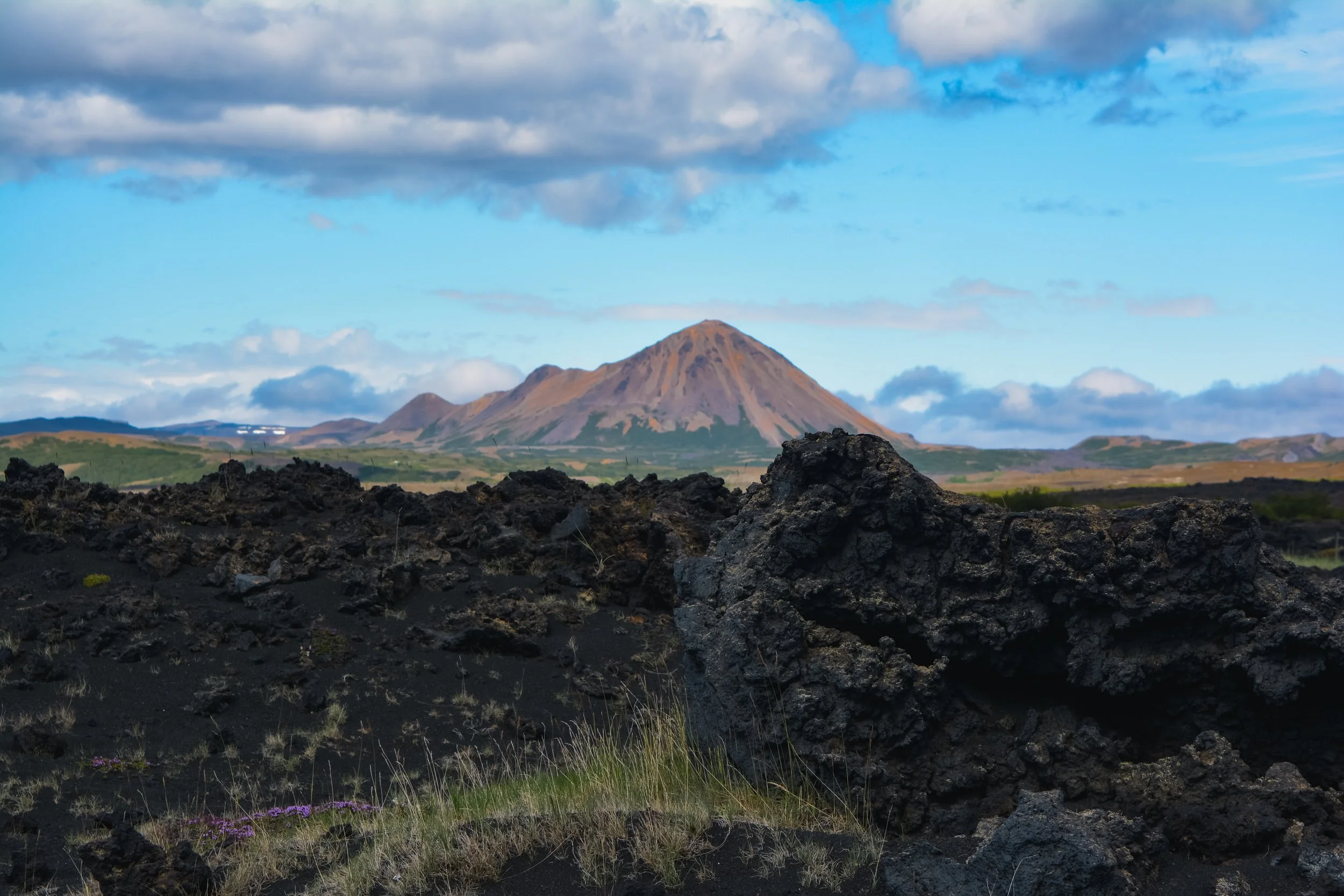 Volcanic landscape with black lava rocks in the foreground, sparse grass, and a distant mountain under a partly cloudy sky.